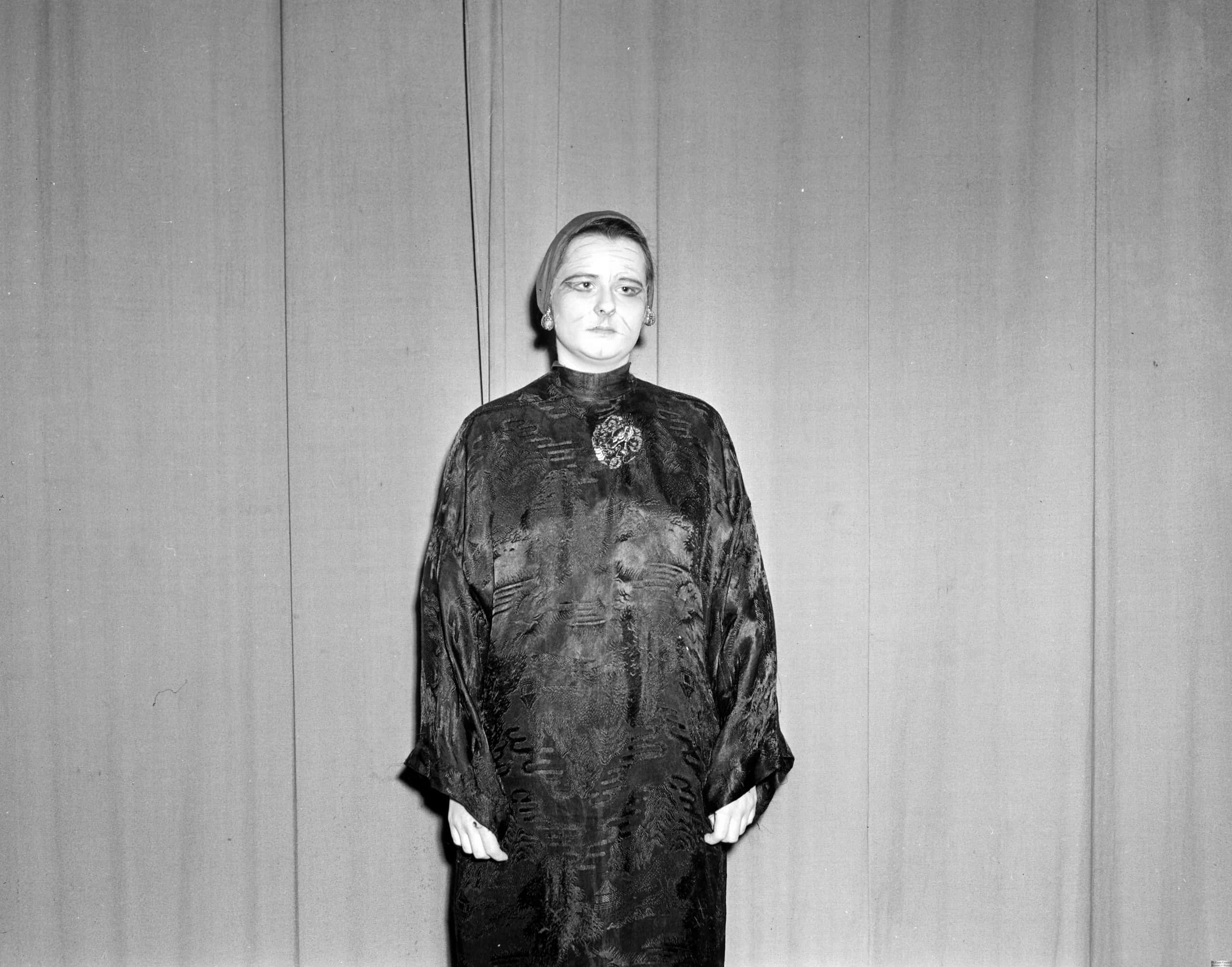 A white actress in yellowface stands in her Dragon Seed costume. She stands with her arms at her sides in front of curtain.
