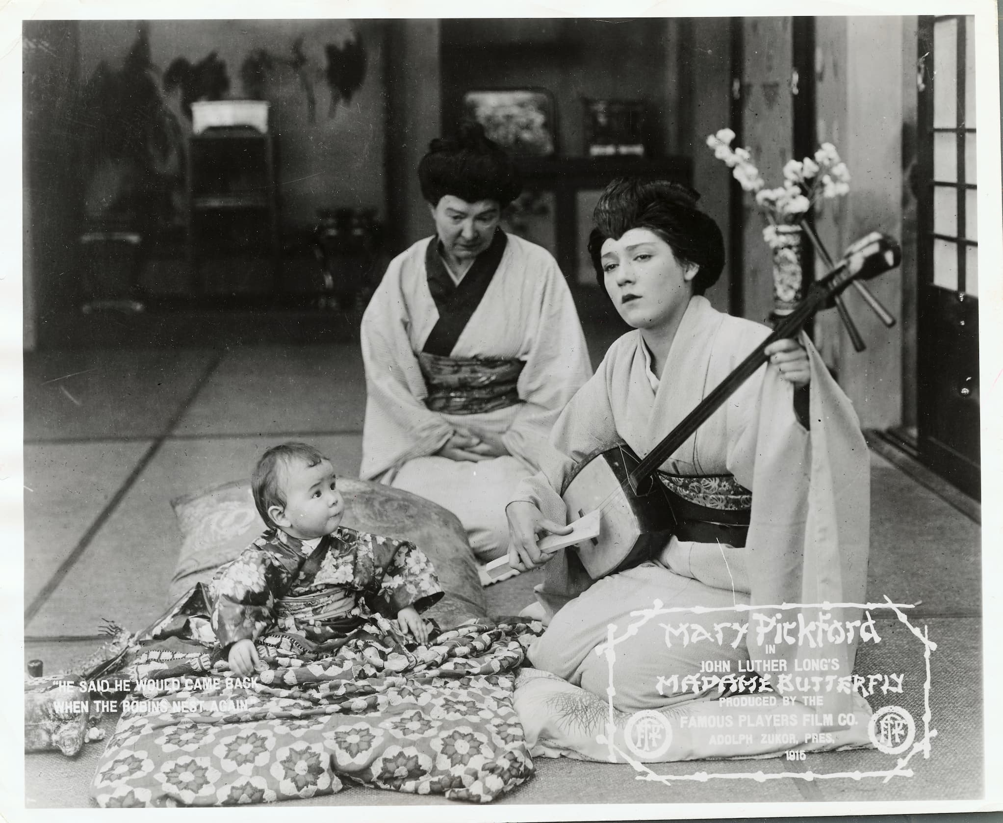 Mary Pickford, in yellowface and dressed in a kimono, plays a three-stringed instrument while kneeling beside a baby.