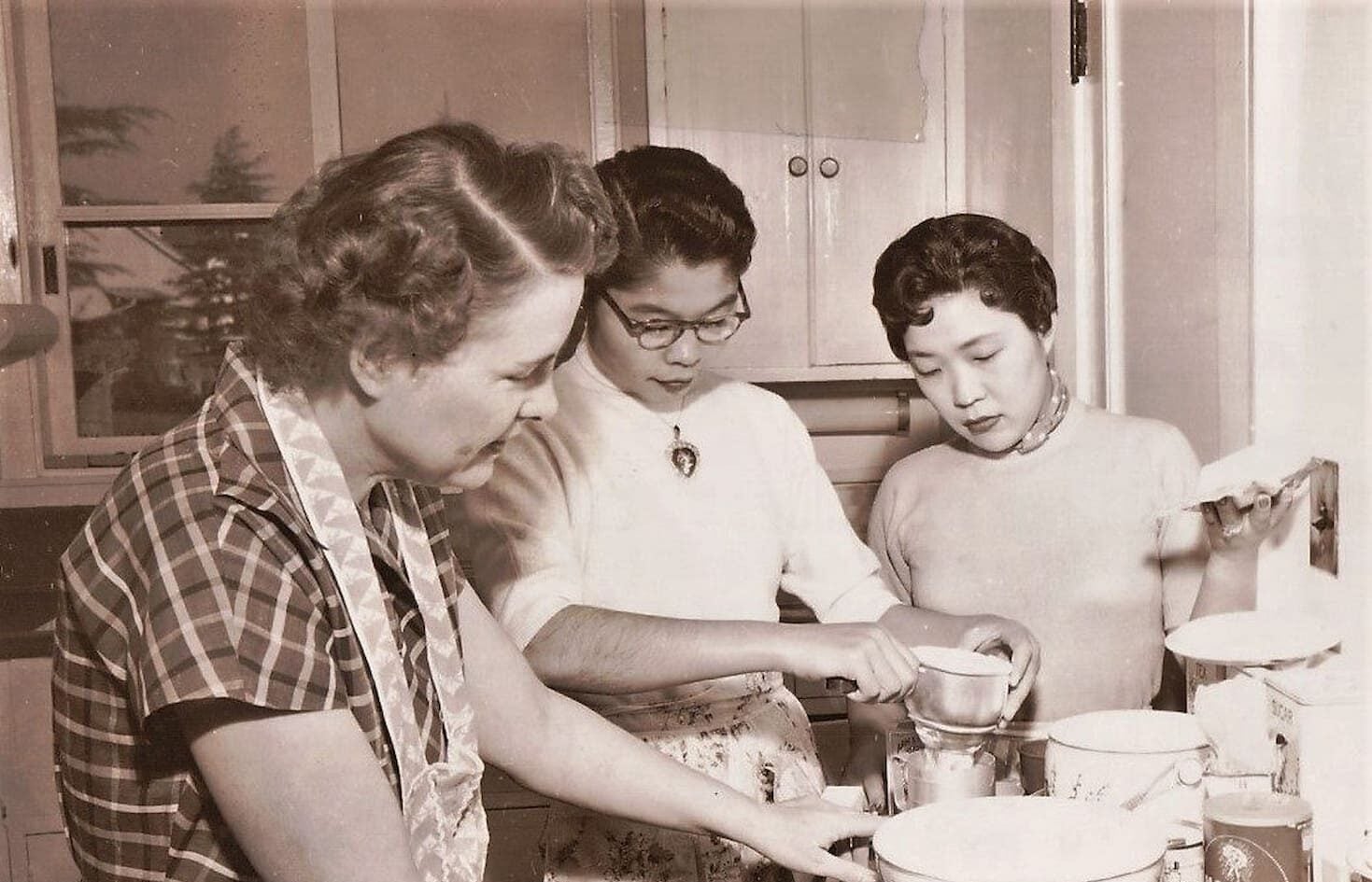 In a kitchen, a white woman wearing an apron and a plaid dress teaches two Japanese women wearing sweaters and floral aprons how to bake.