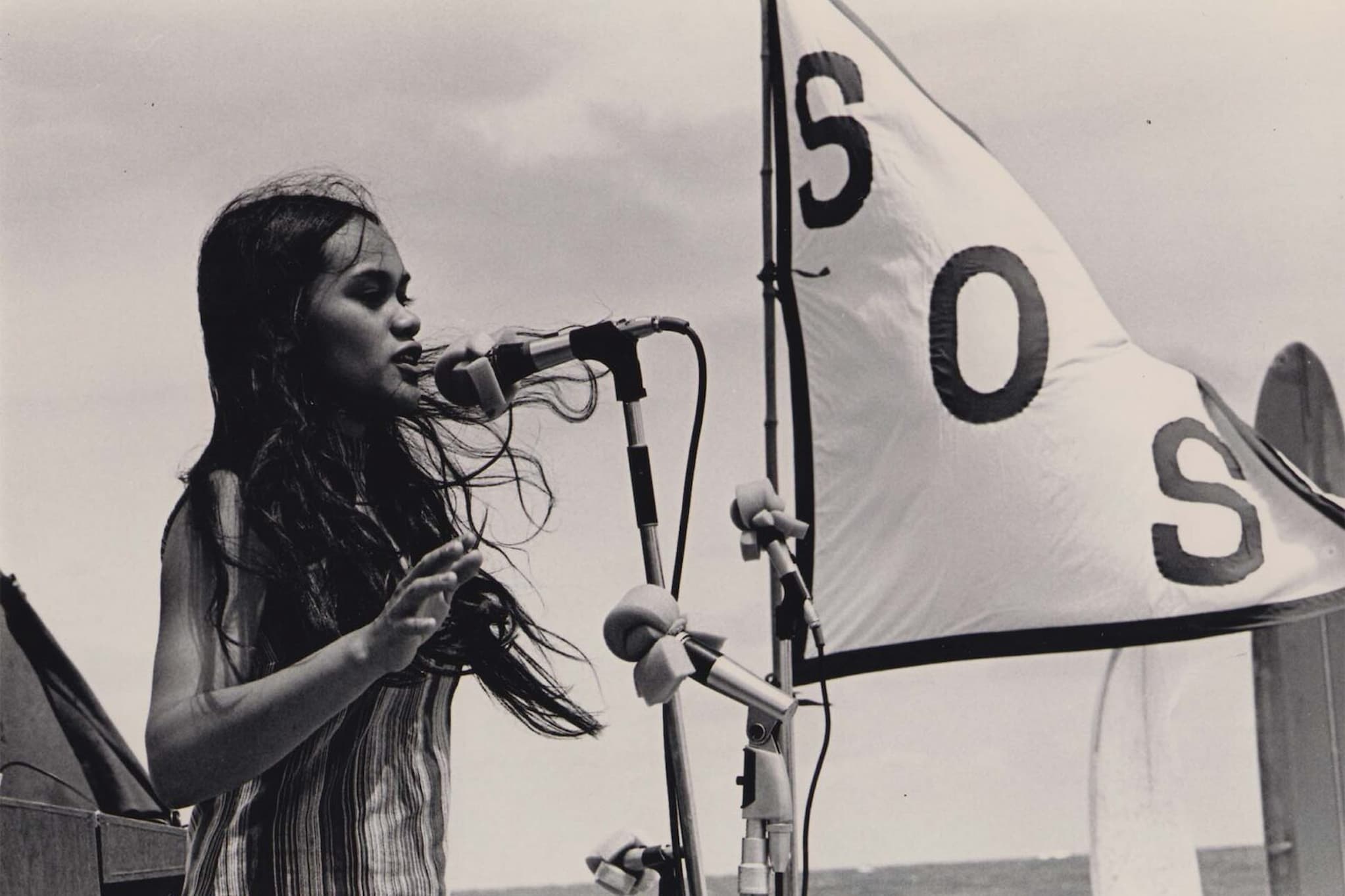 Kanaka ʻŌiwi activist Terrilee Kekoʻolani-Raymond speaks into a microphone during outdoor rally. Next to her is a white flag embroidered with "SOS."