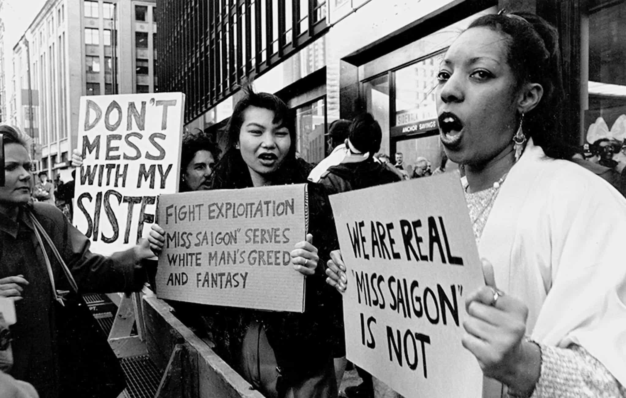 Protestors behind barricade carry signs protesting Miss Saigon as they line sidewalk. Opposite them a woman reads the signs.
