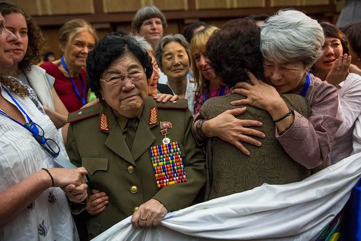 Two elderly women hug while an elderly woman wearing a decorated military uniform grasps white quilt with her hand. Smiling women surround them.