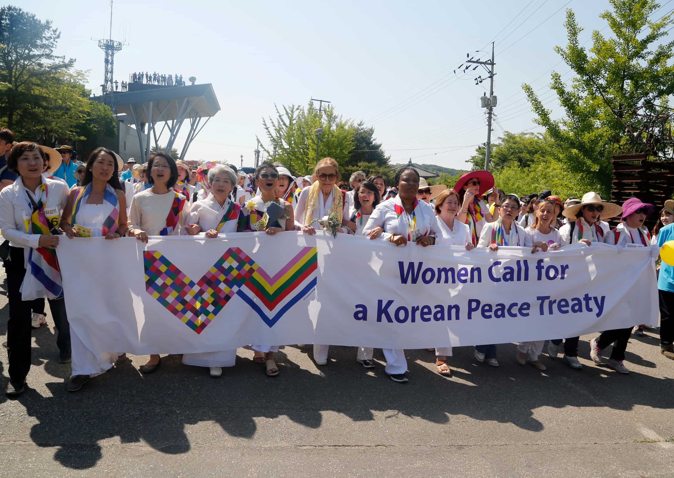 Members of Women Cross DMZ hold a banner that reads "Women Call for a Korean Peace Treaty" with a stylized "w" logo as they march outside.