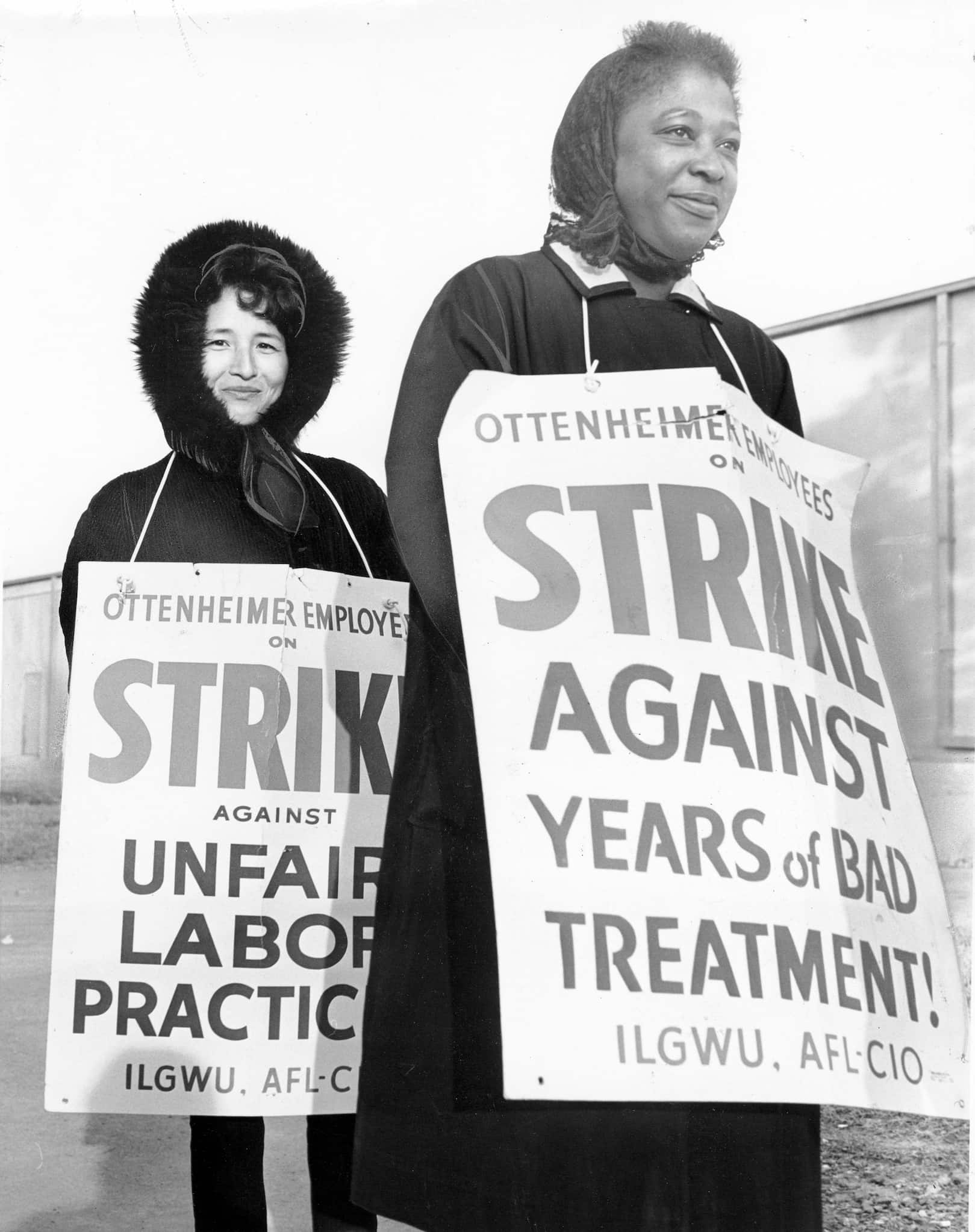 An Asian woman and a Black woman of the International Ladies' Garment Workers Union stand with signs around their necks explaining reasons to strike.