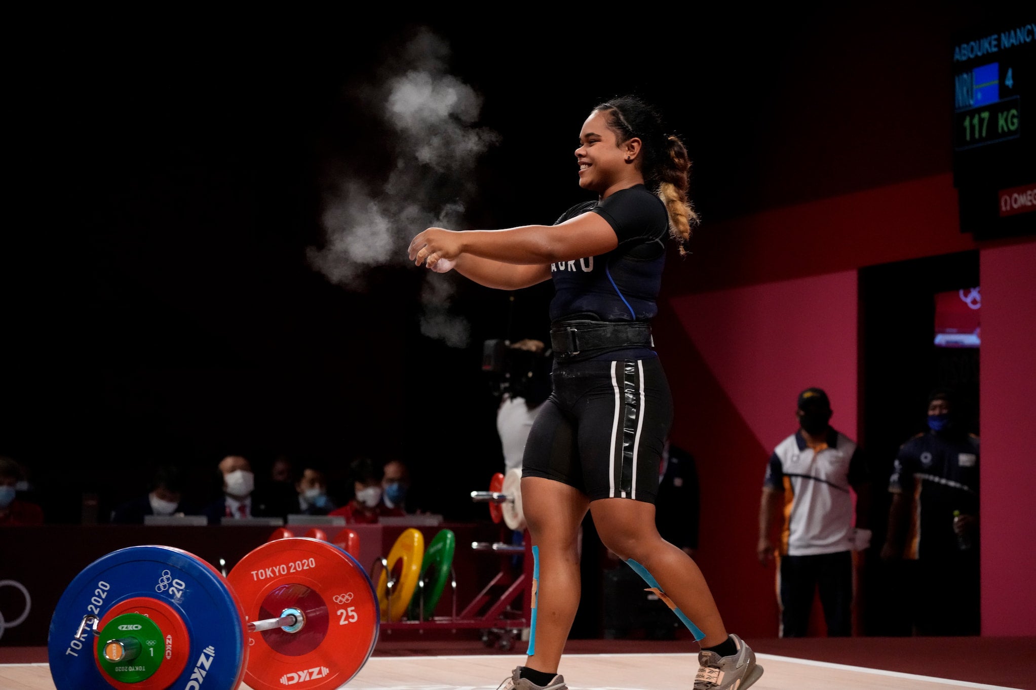 Nancy Abouke, dressed in a weightlifting uniform, claps her chalk-filled hands in front of a deadlift bar. Clouds of chalk float above her hands.