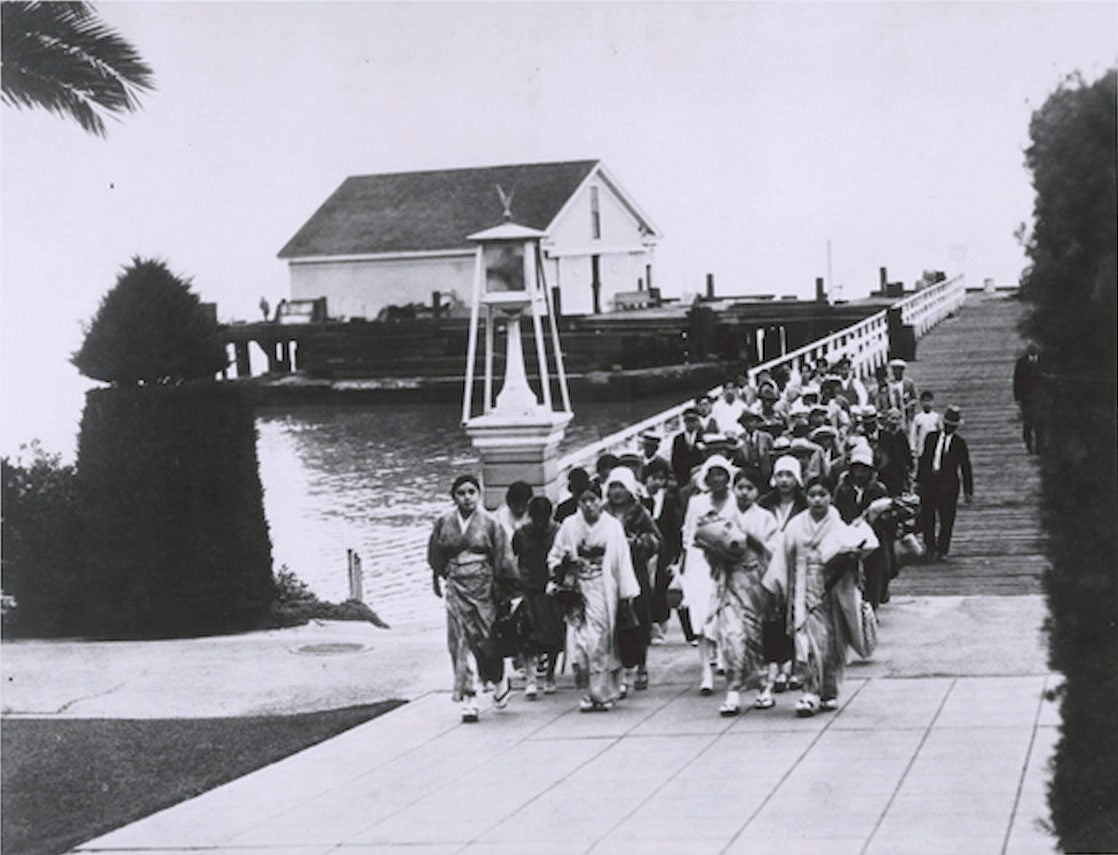 A crowd of Japanese immigrants walk along wide, planked pier towards Angel Island. Behind them is a large building at edge of pier.