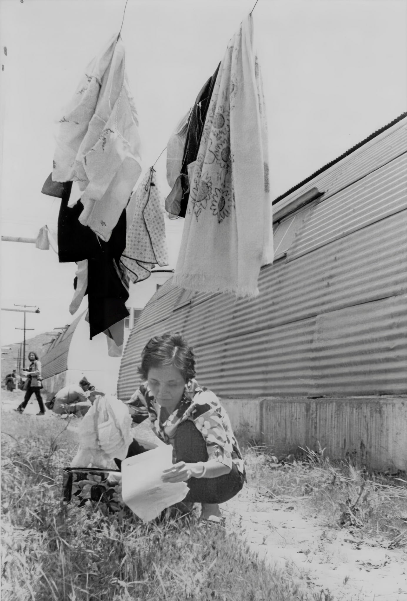 Black-and-white photo of a woman hunched over in the grass holding a bag and some papers. Above her, clothes dry on a clothesline.