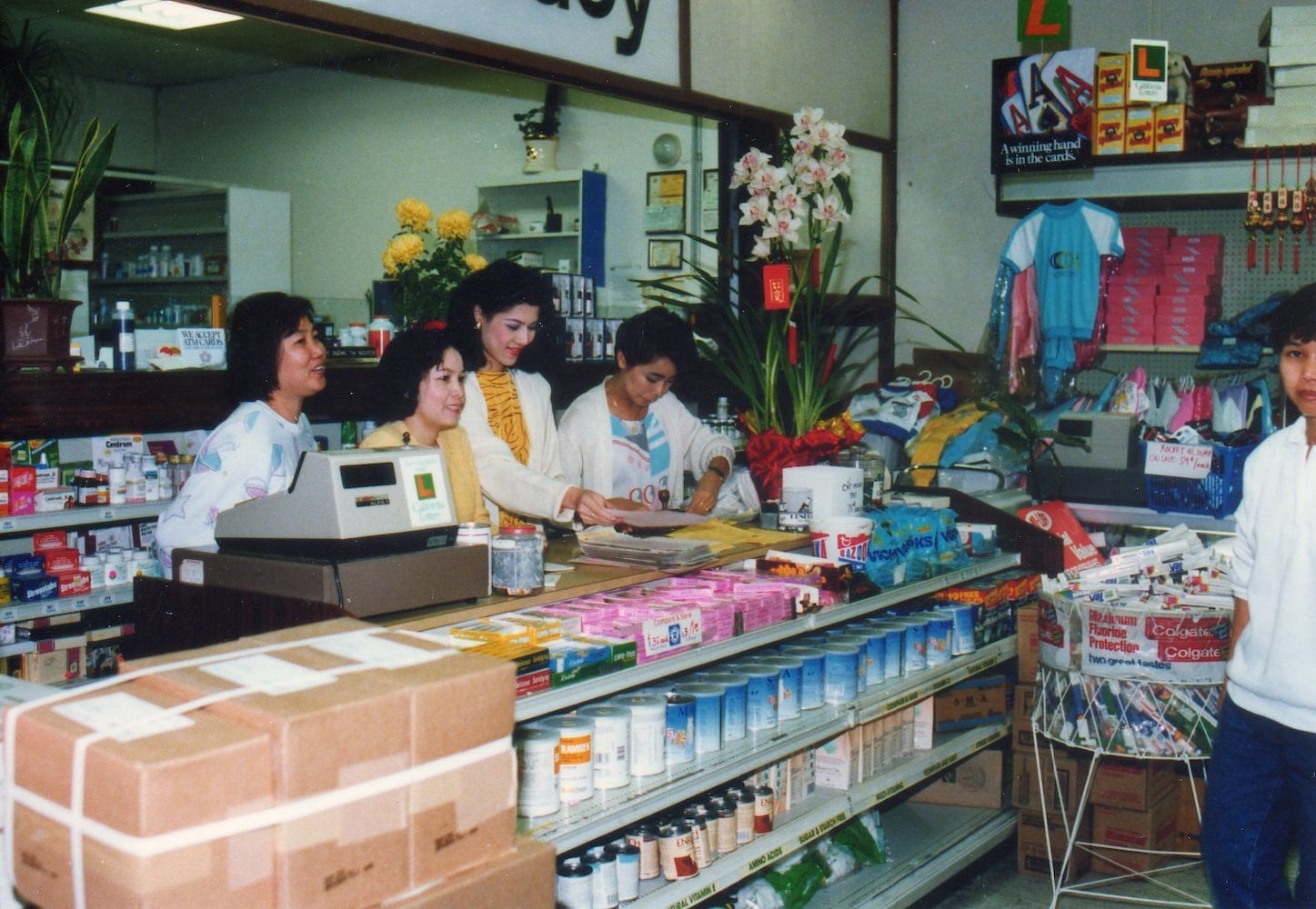 Four women smiling and talking stand behind a cashier counter in a pharmacy. A man stands to the right looking at the camera.