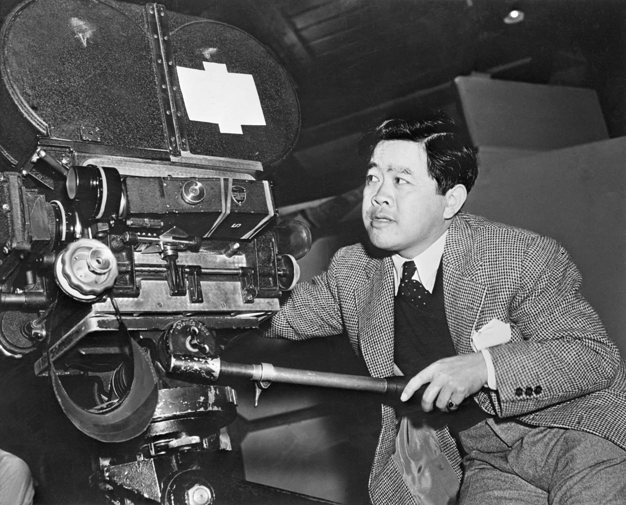 James Wong Howe, a Chinese American cinematographer dressed in a tweed jacket, vest, pants, shirt and tie, sits behind a movie camera.