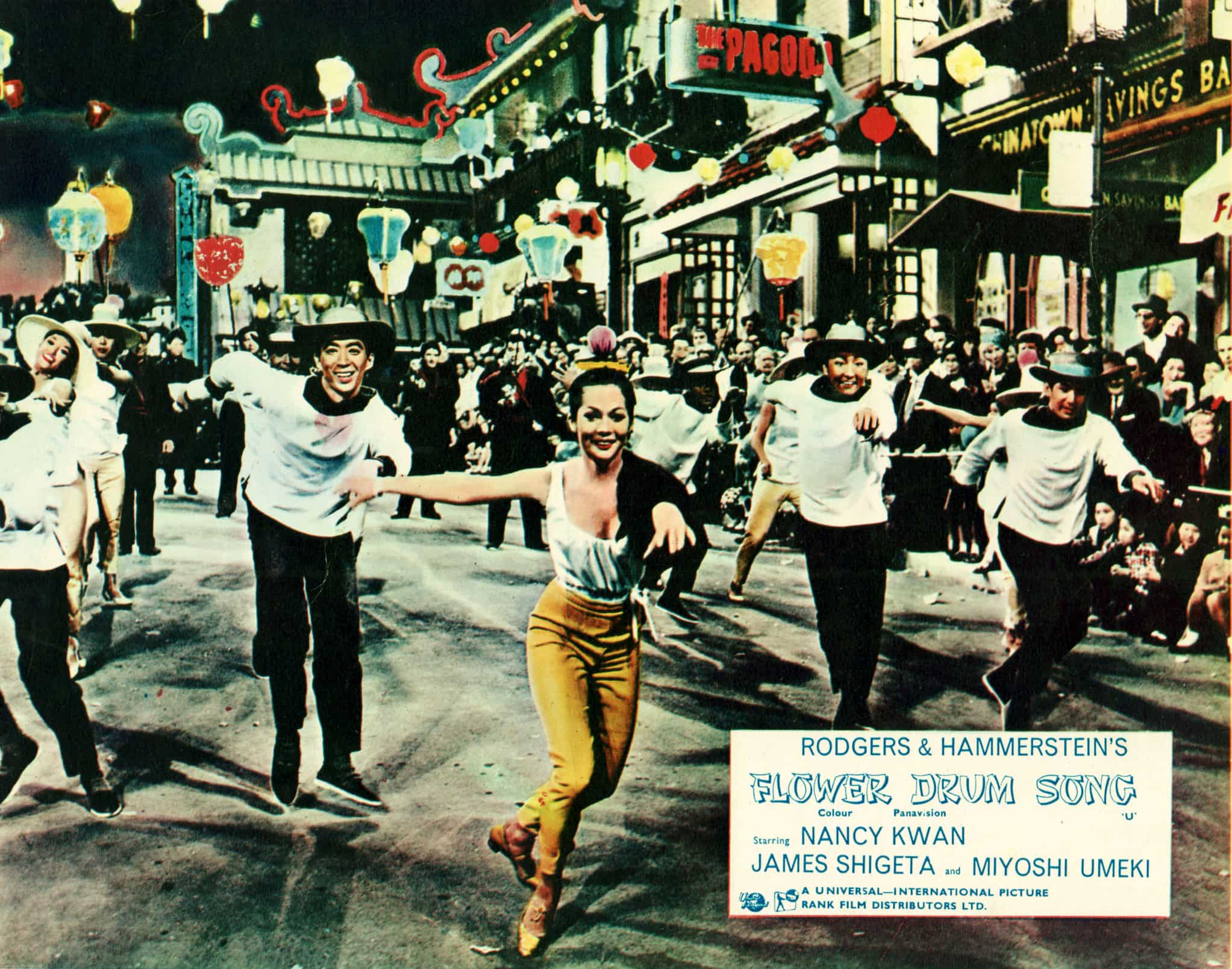 A still from Flower Drum Song shows a crowd gathered in a Chinatown street to watch a parade. Several smiling performers dance in the street.