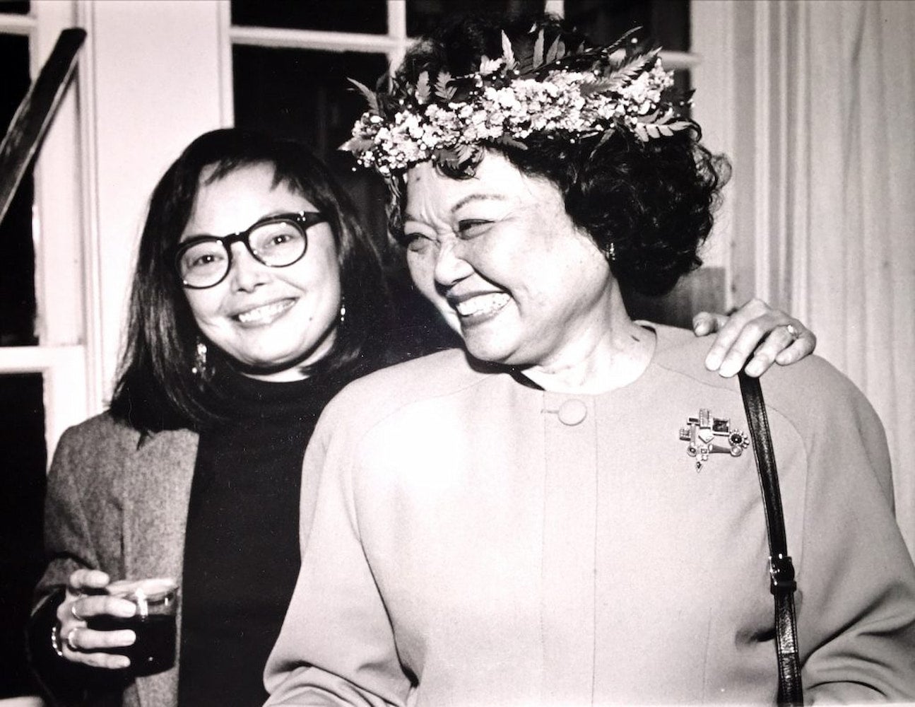 Gwendolyn Mink, woman with glasses, smiles with her arm around mother, Patsy Mink, woman wearing a lei poʻo and brooch, in indoor candid.