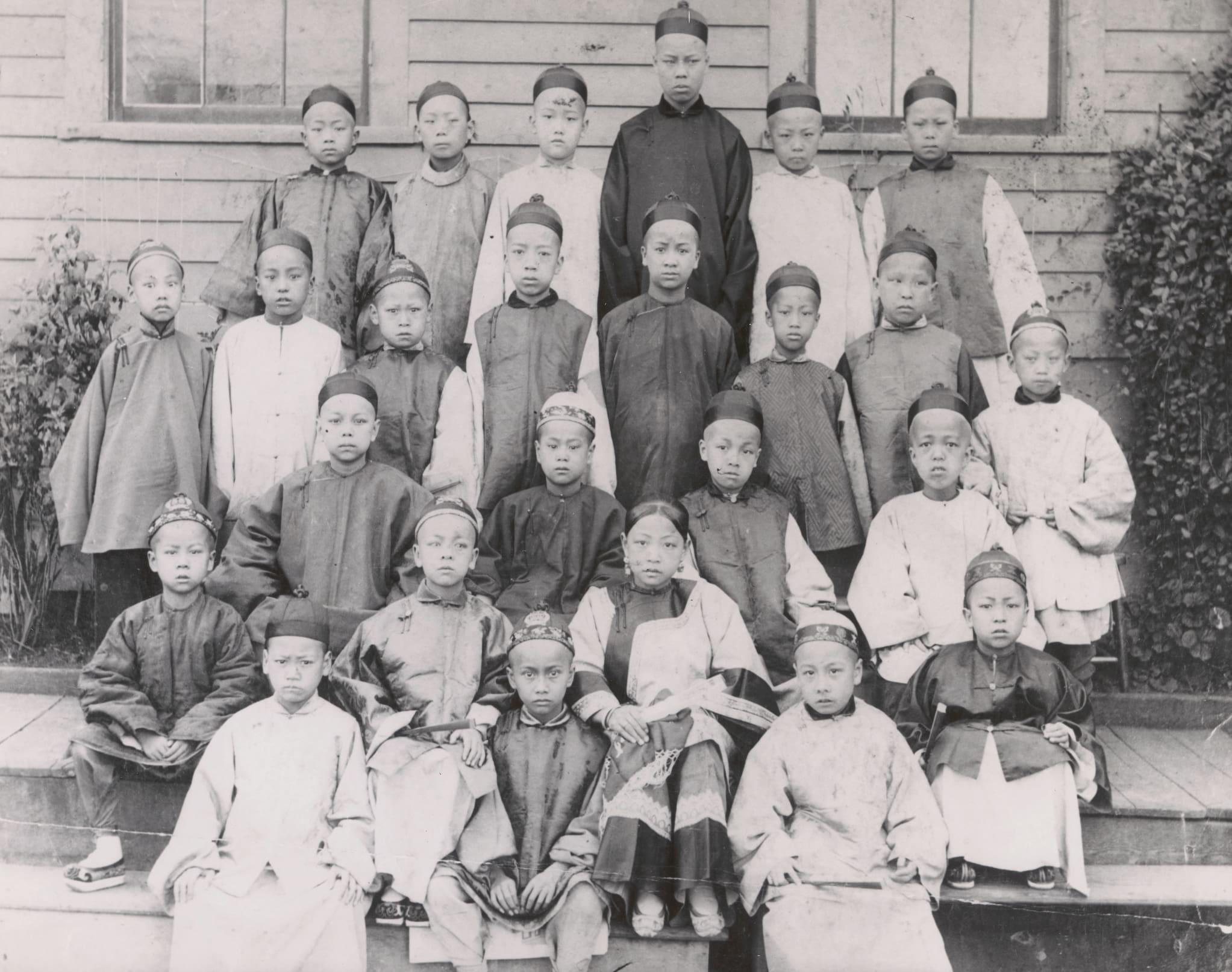 Chinese school students dressed in traditional Chinese clothing stand for a class photo. Mamie and Frank Tape are seen in the center second row.
