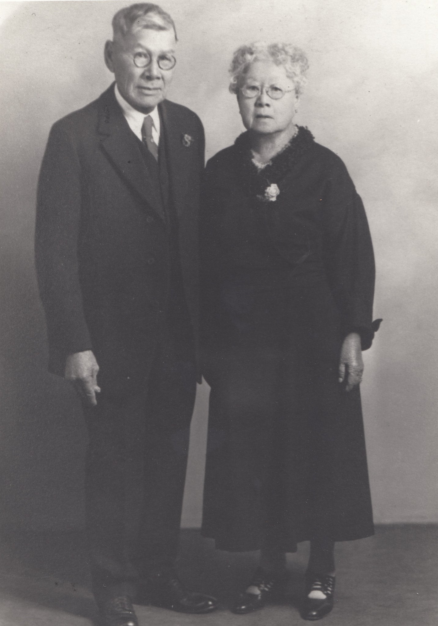 Mary Tape in a black dress and round glasses and Joseph Tape in a black suit and round glasses pose for a portrait in black and white.