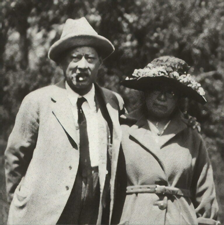 Frank Tape in a tan coat and hat stands to the left of Mary Tape in a tan coat and sun hat pose for a photo.