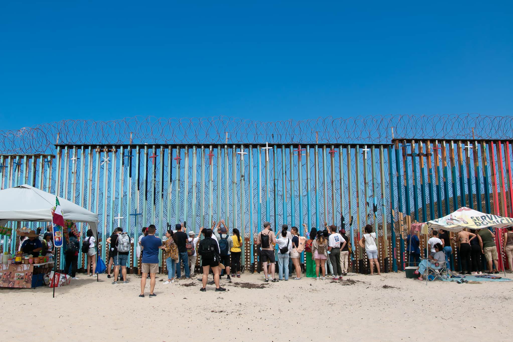Union leaders, community organizers, and worker activists visit the wall at the U.S.-Mexico border. The wall is topped with barbed wire and crosses.