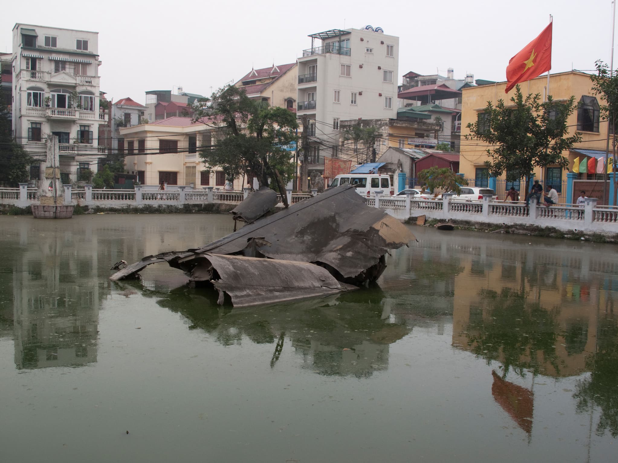 In Hữu Tiệp Lake, the wreckage of a B-52 bomber is half-submerged. Around the lake are buildings and a Vietnamese flag fluttering in the wind.