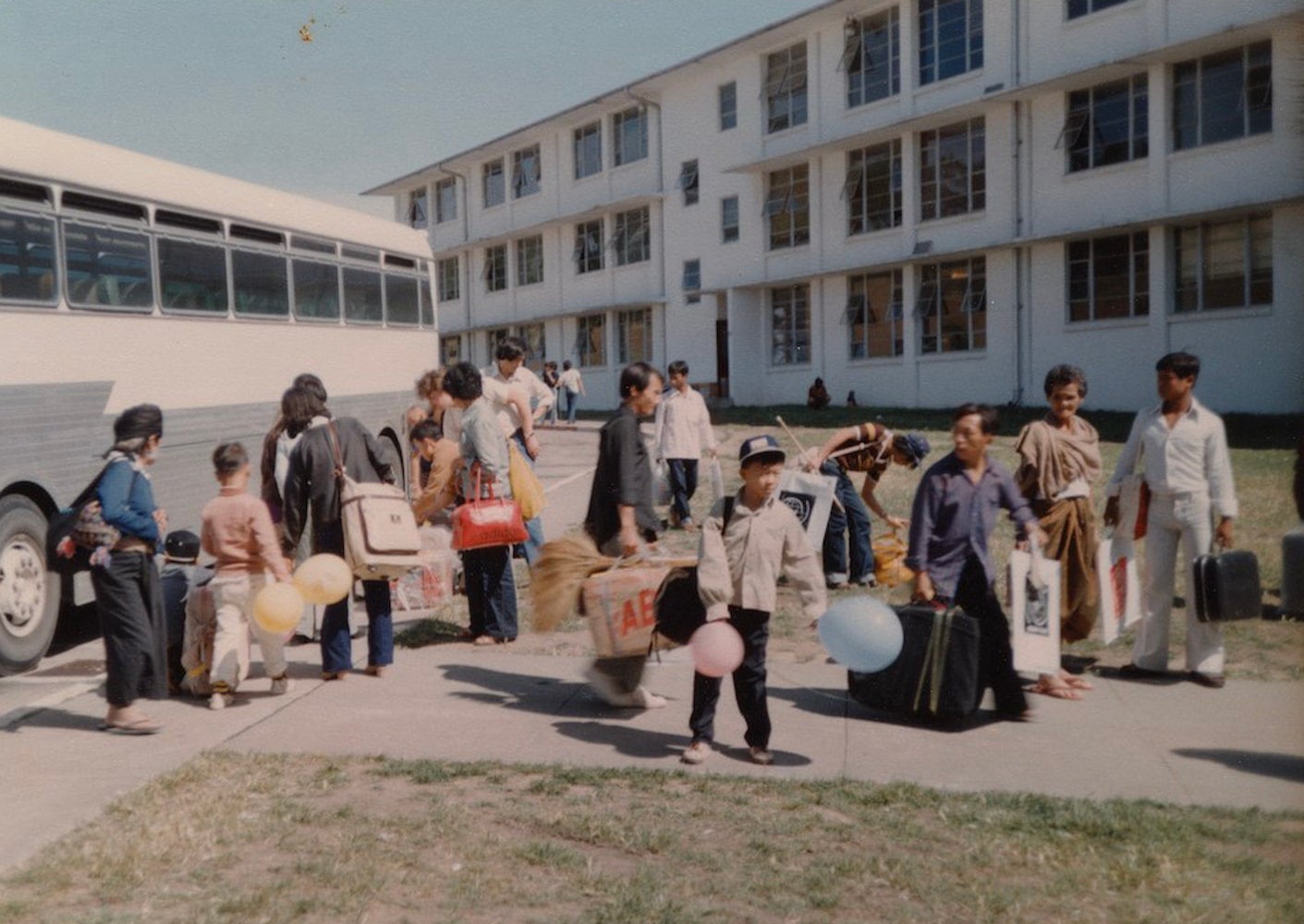 Laotian refugees debark from a bus parked in front of a building at Hamilton Air Force Base. They carry their belongings.