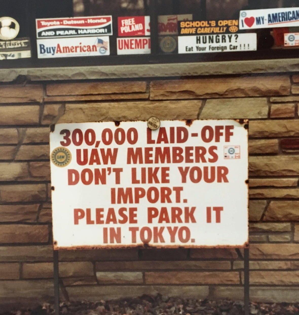 White sign with red text reads, "300,000 Laid-Off UAW Members Don't Like Your Import. Please Park It In Tokyo." Bumper stickers surround sign.