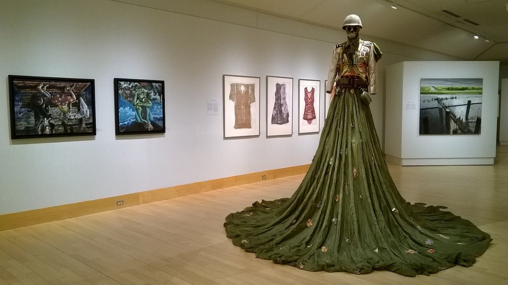 Art piece of a statue with a skull head wearing a soldier's helmet and a dress made out of a military parachute displayed in a gallery.