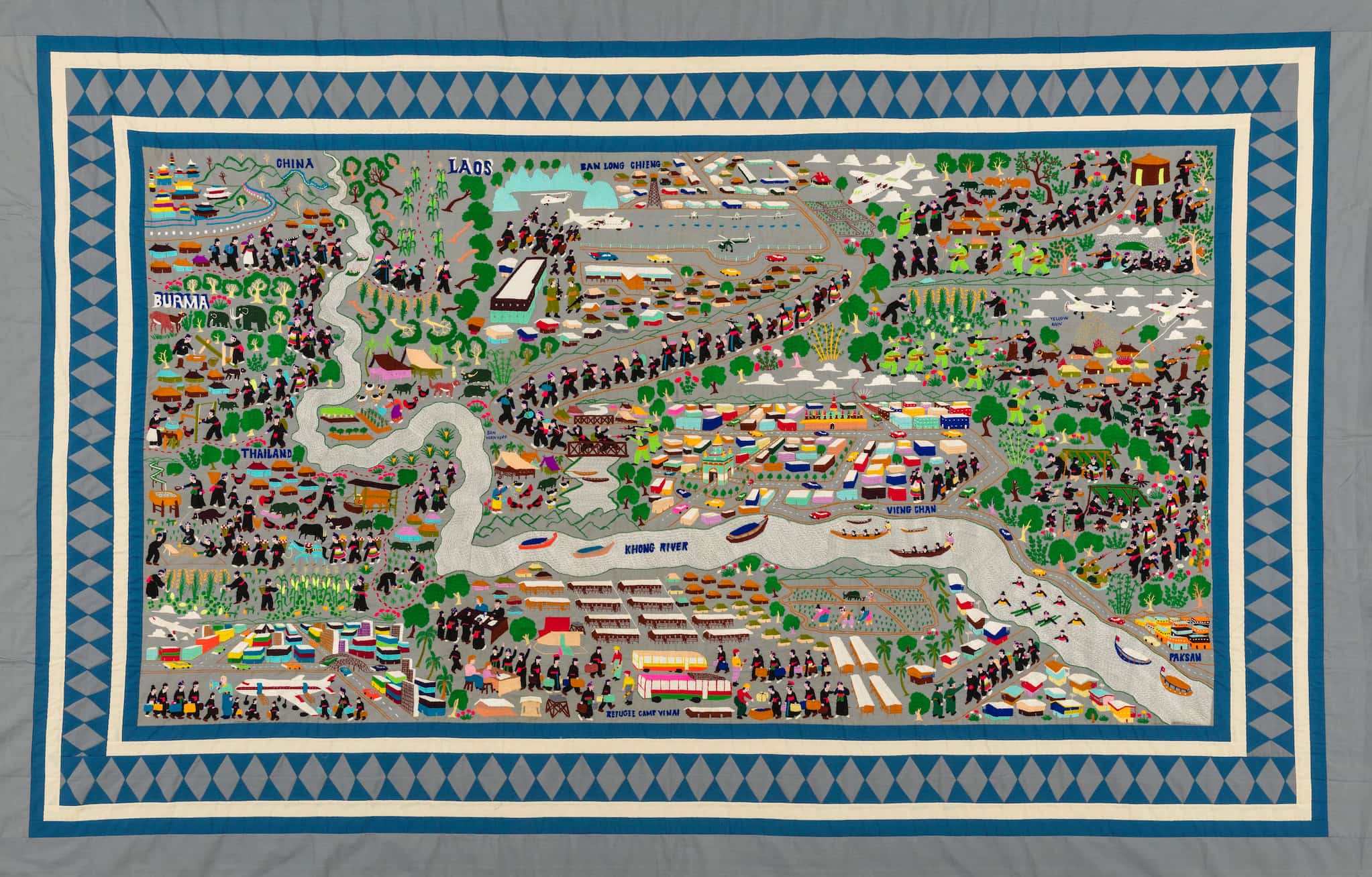 Set against gray fabric, this detailed story cloth shows Hmong people as they walk through lands with colorful buildings bisected by long river.