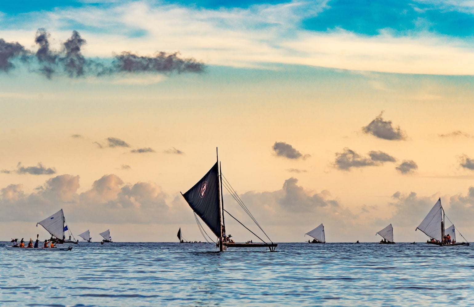 Unfurled sails from sakmans fill the horizon. Canoes sit on the calm ocean along with the sakmans. Two birds fly over the sakmans in a cloudy sky.