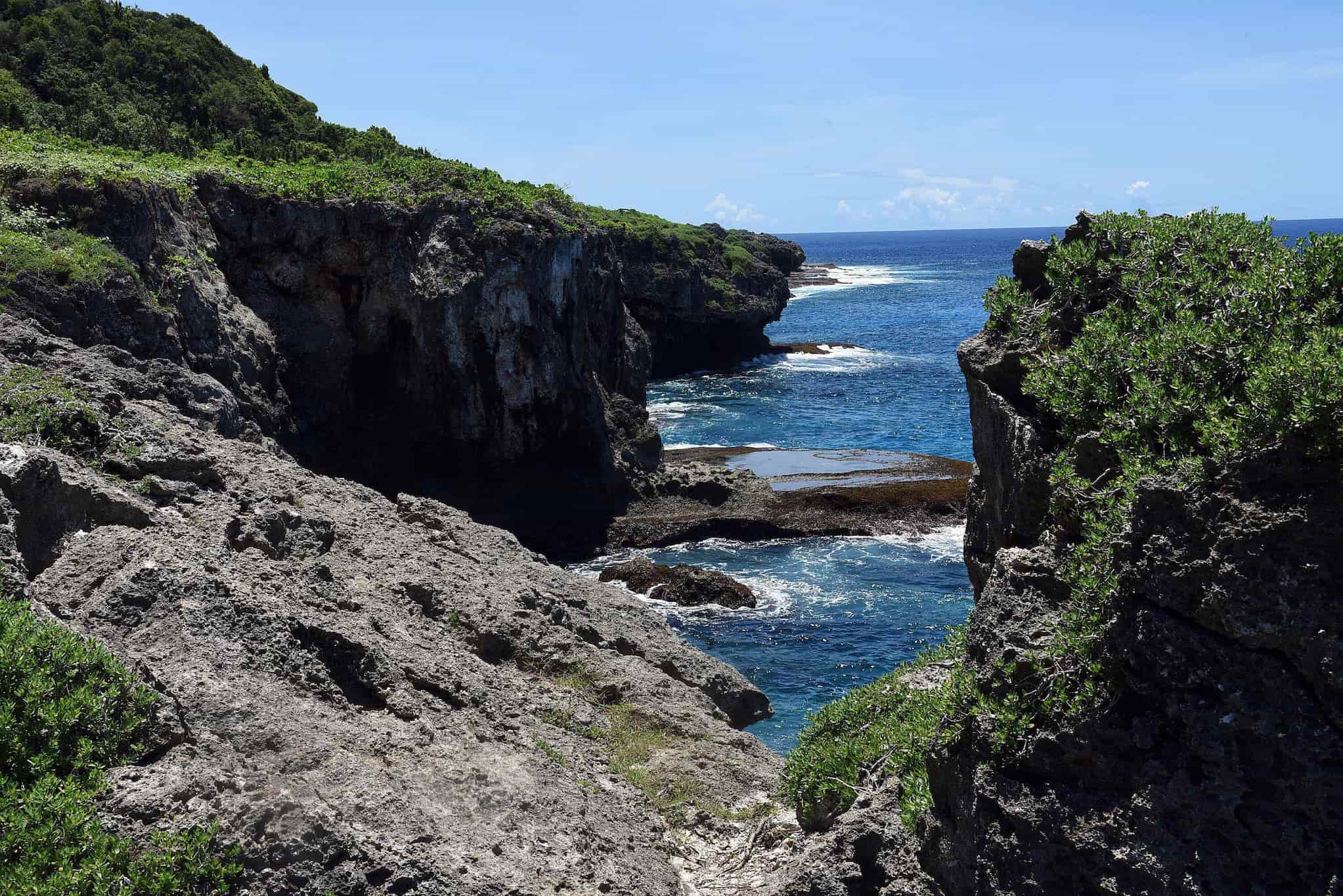 The coast of Pågat on a sunny day. Clear, blue water is surrounded by limestone cliffs topped with dense, green foliage.