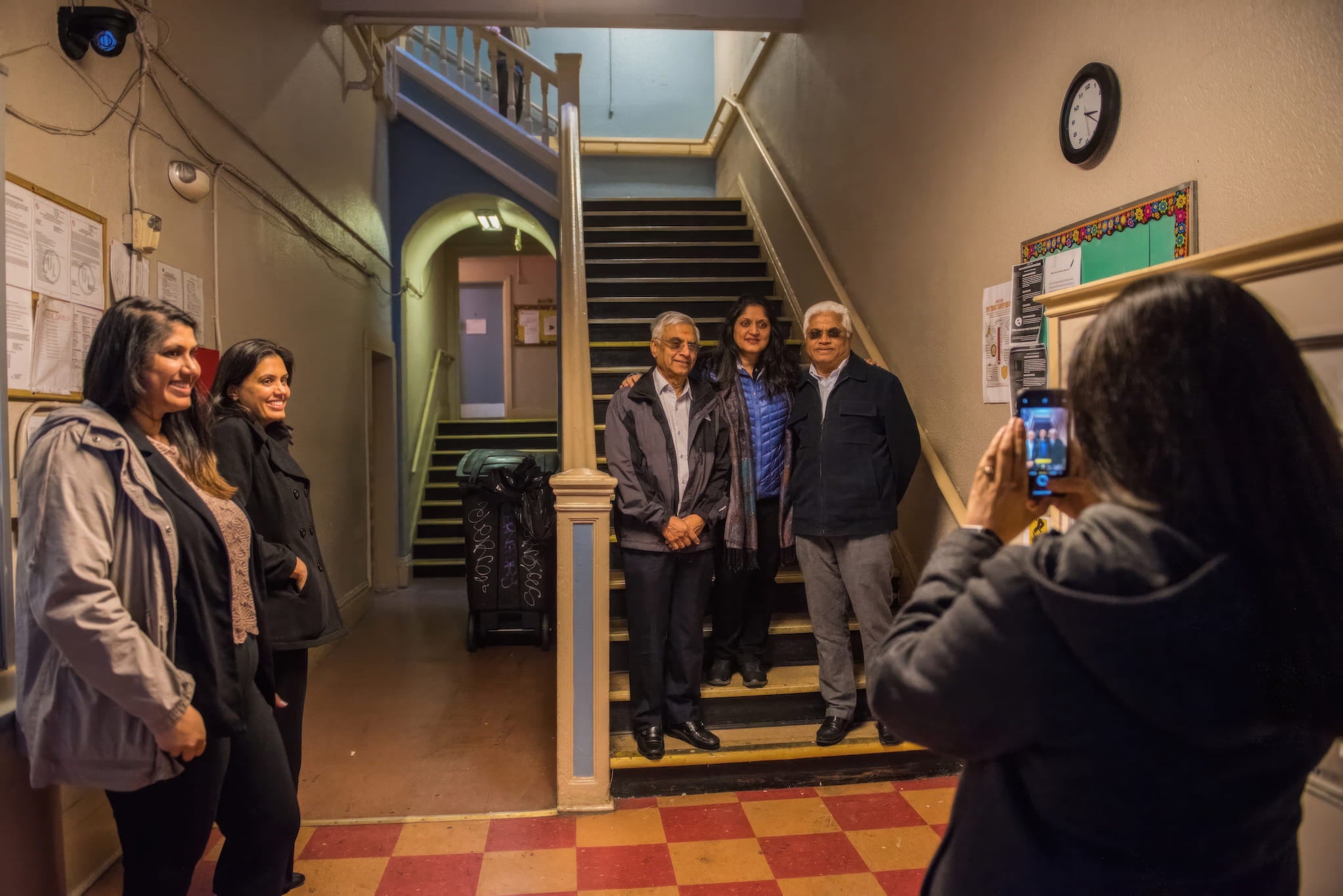 Pratima Patel takes a photo of her family members standing at the bottom of their hotel's staircase. Her sisters watch, standing off to the side.