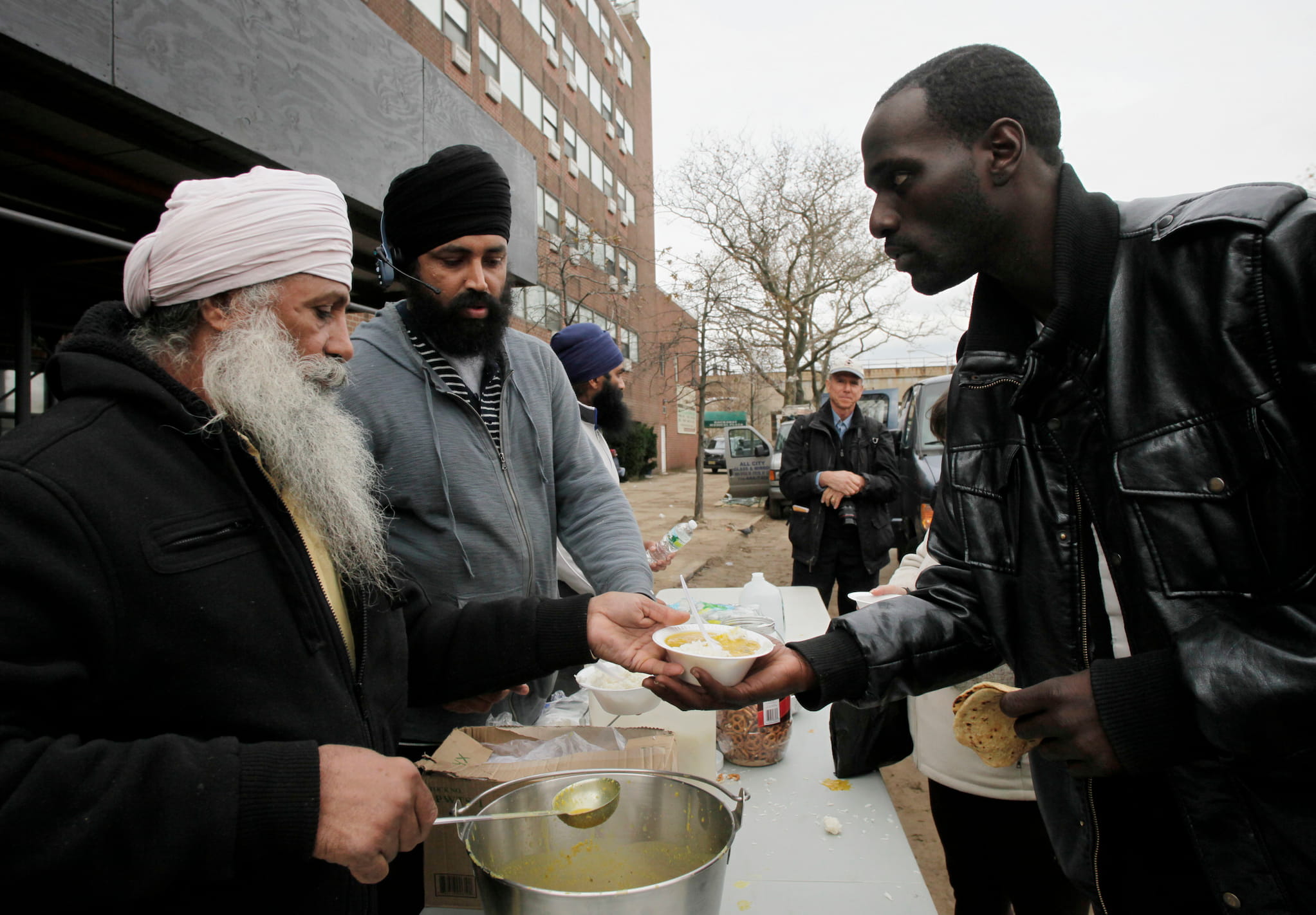 After Hurricane Sandy, Sikh volunteers dressed in hoodies and turbans serve a hot meal to a Black man wearing a leather jacket.
