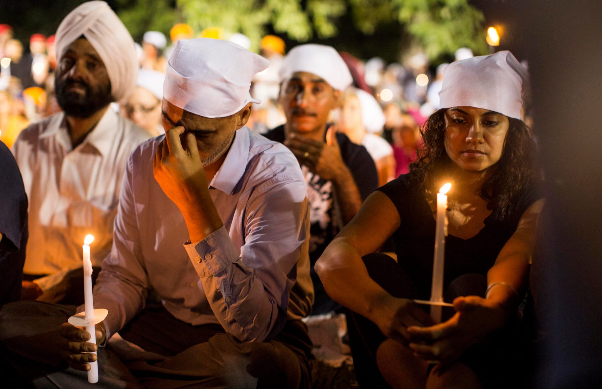 At an evening candlelight vigil for the victims of a mass shooting at the Sikh Temple, seated mourners wear white head wraps and hold candles.
