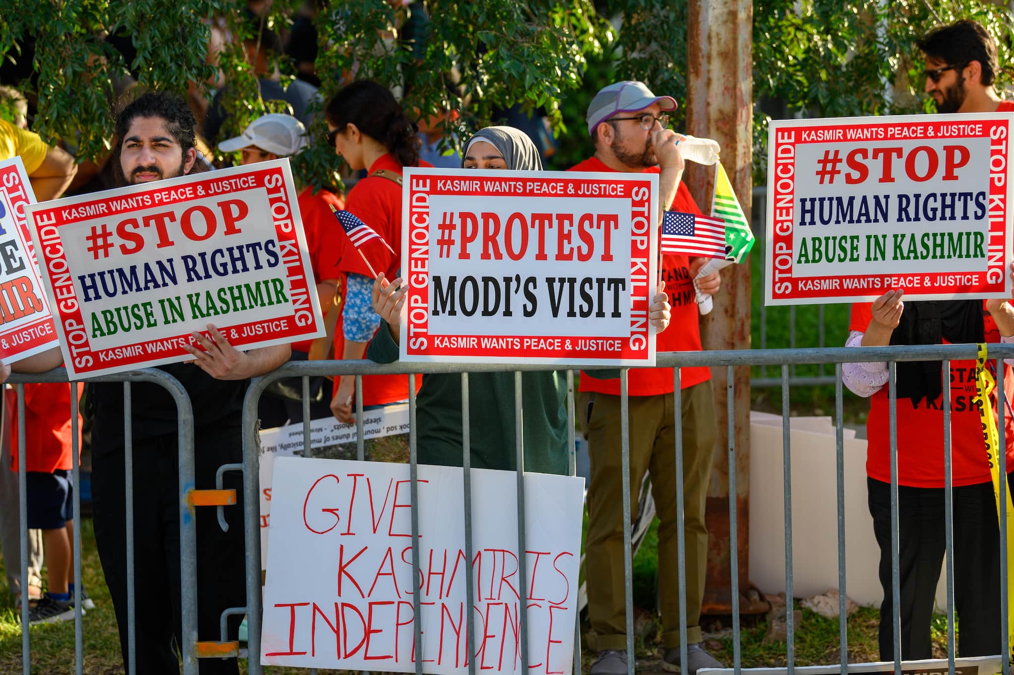 Demonstrators, wearing red t-shirts, stand behind metal barricades with signs protesting the Indian government and Modi's visit to Texas.