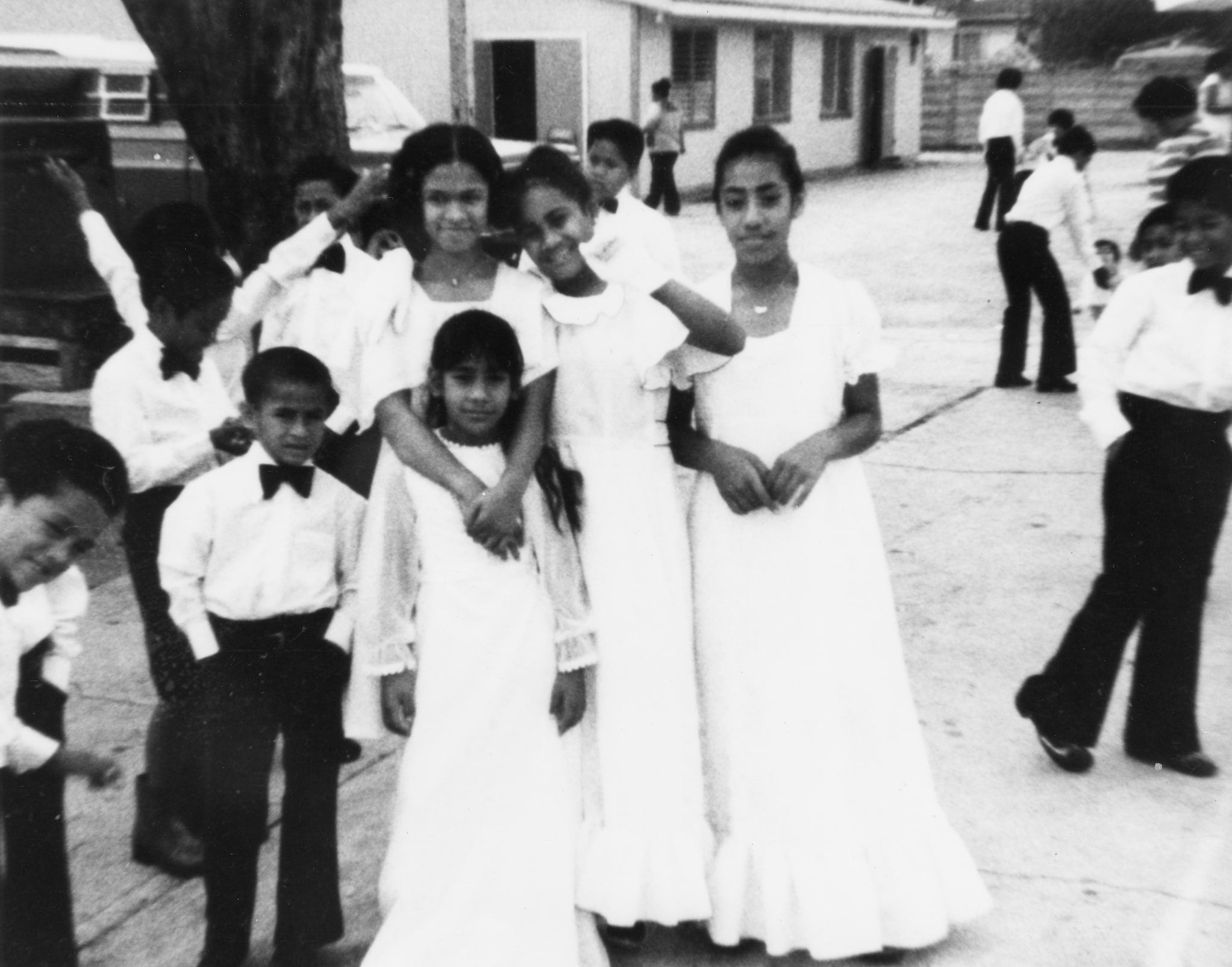 Four Samoan girls and one Samoan boy pose and wear white outside of a church during White Sunday. Other similarly dressed children socialize around them.