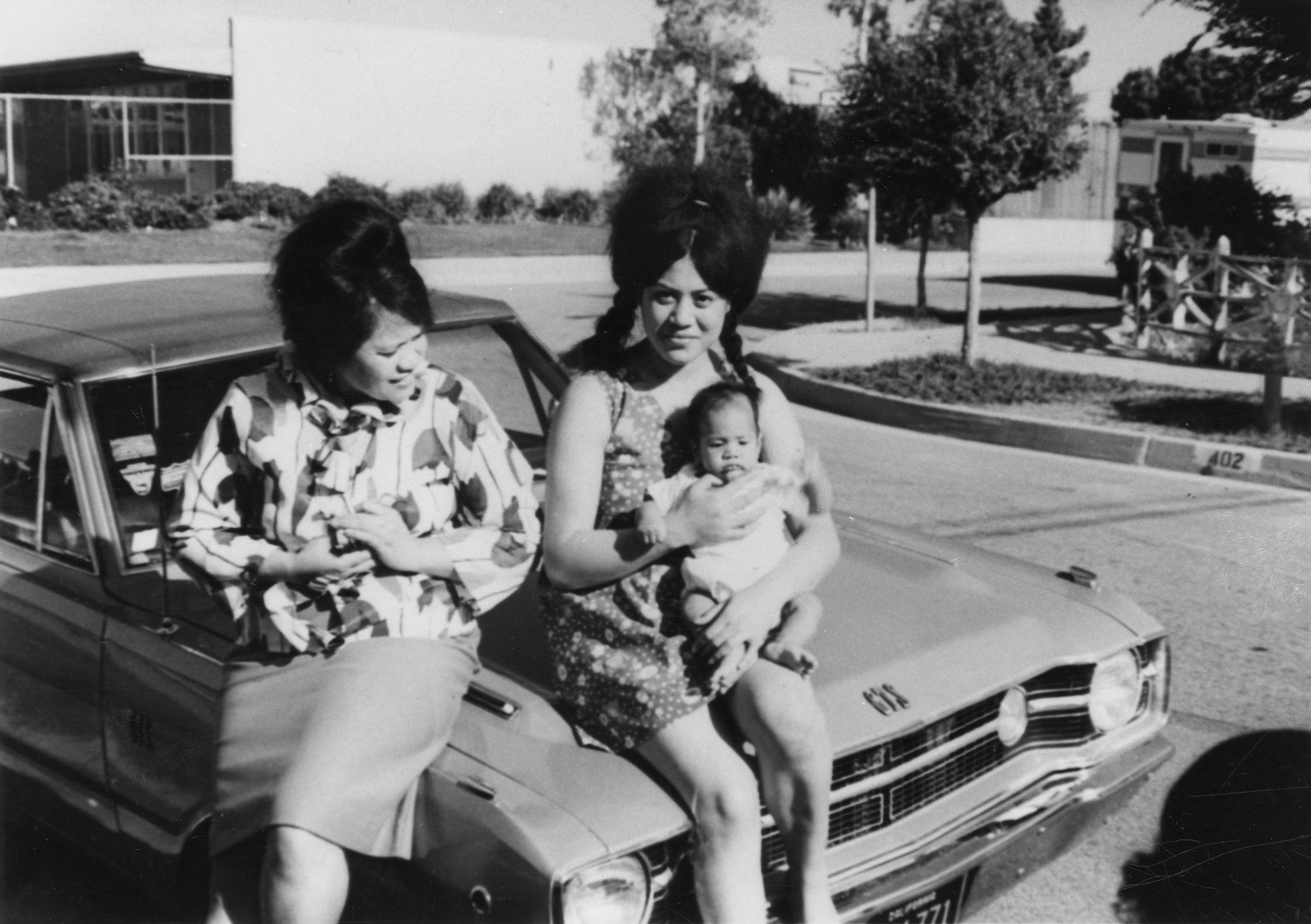 Samoan American Liz sits on the hood of a car while holding her baby. Her cousin Vaisola sits next to her and leans over to see the baby.