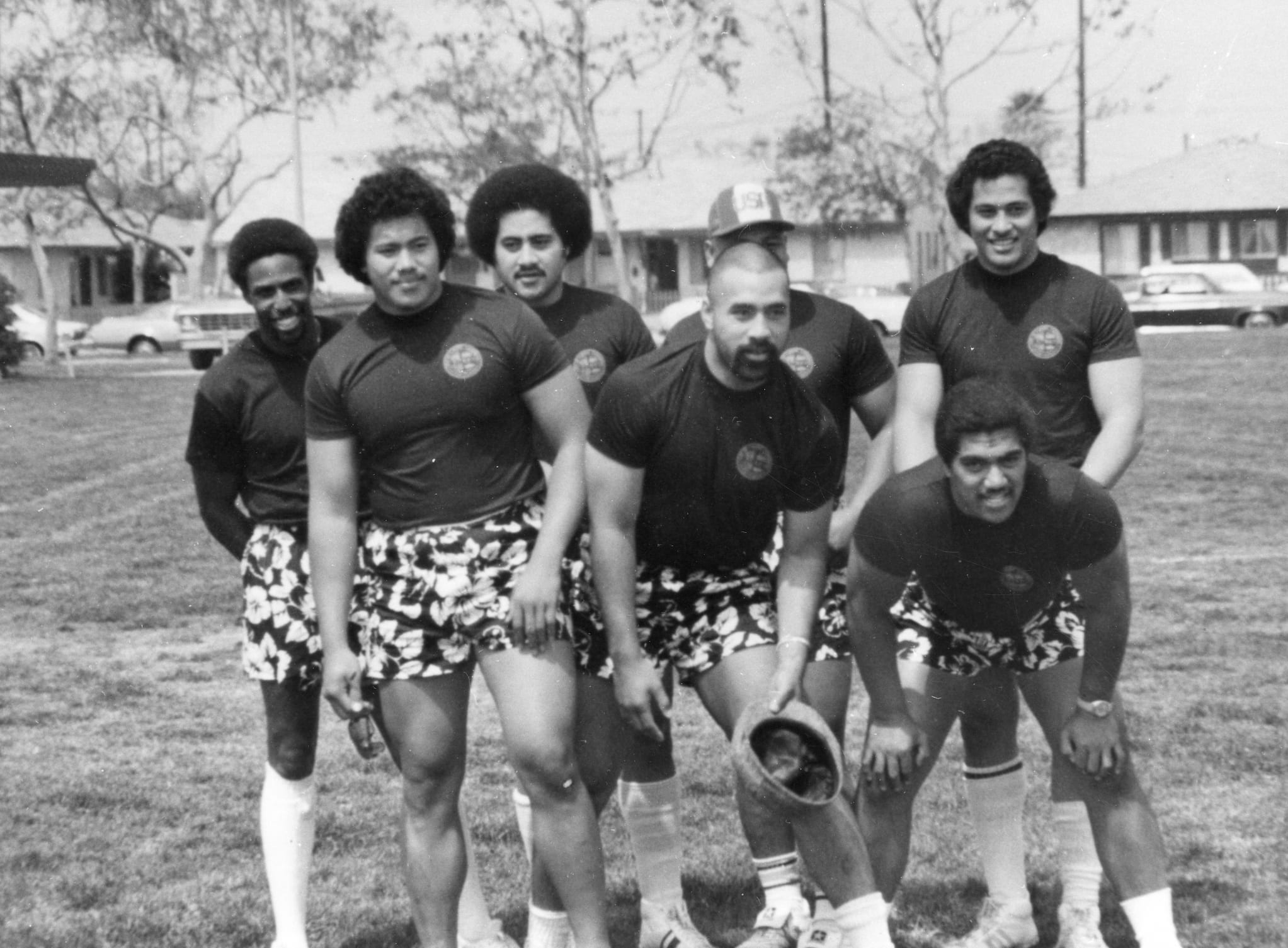 Group photo of seven Samoan American football players on the field at Scott Park. They are dressed in matching t-shirts and hibiscus print shorts.