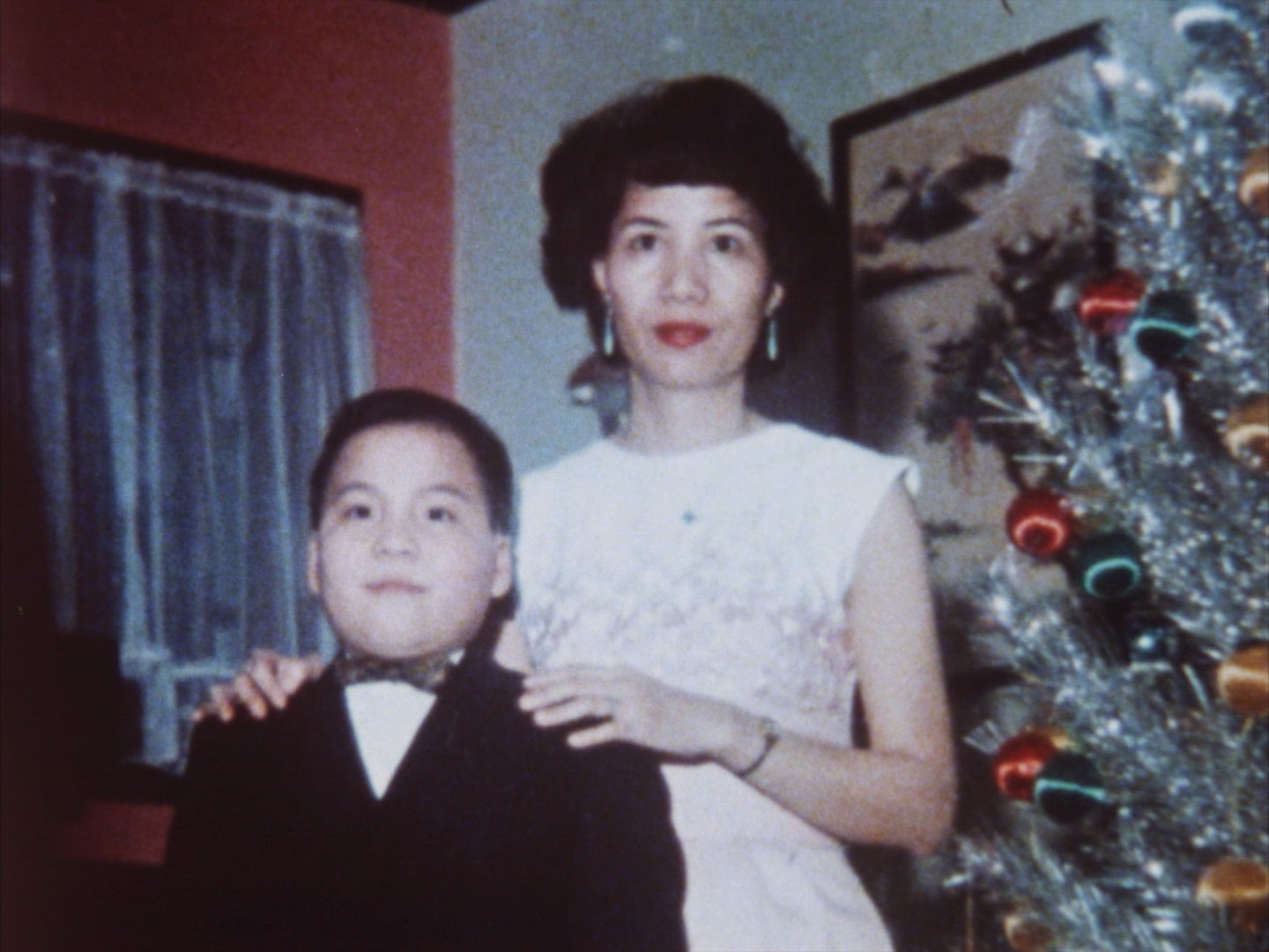 Vincent Chin, a young boy dressed in suit jacket and bowtie, poses next to Christmas tree with his mother, Lily Chin. Her hands are on his shoulders.