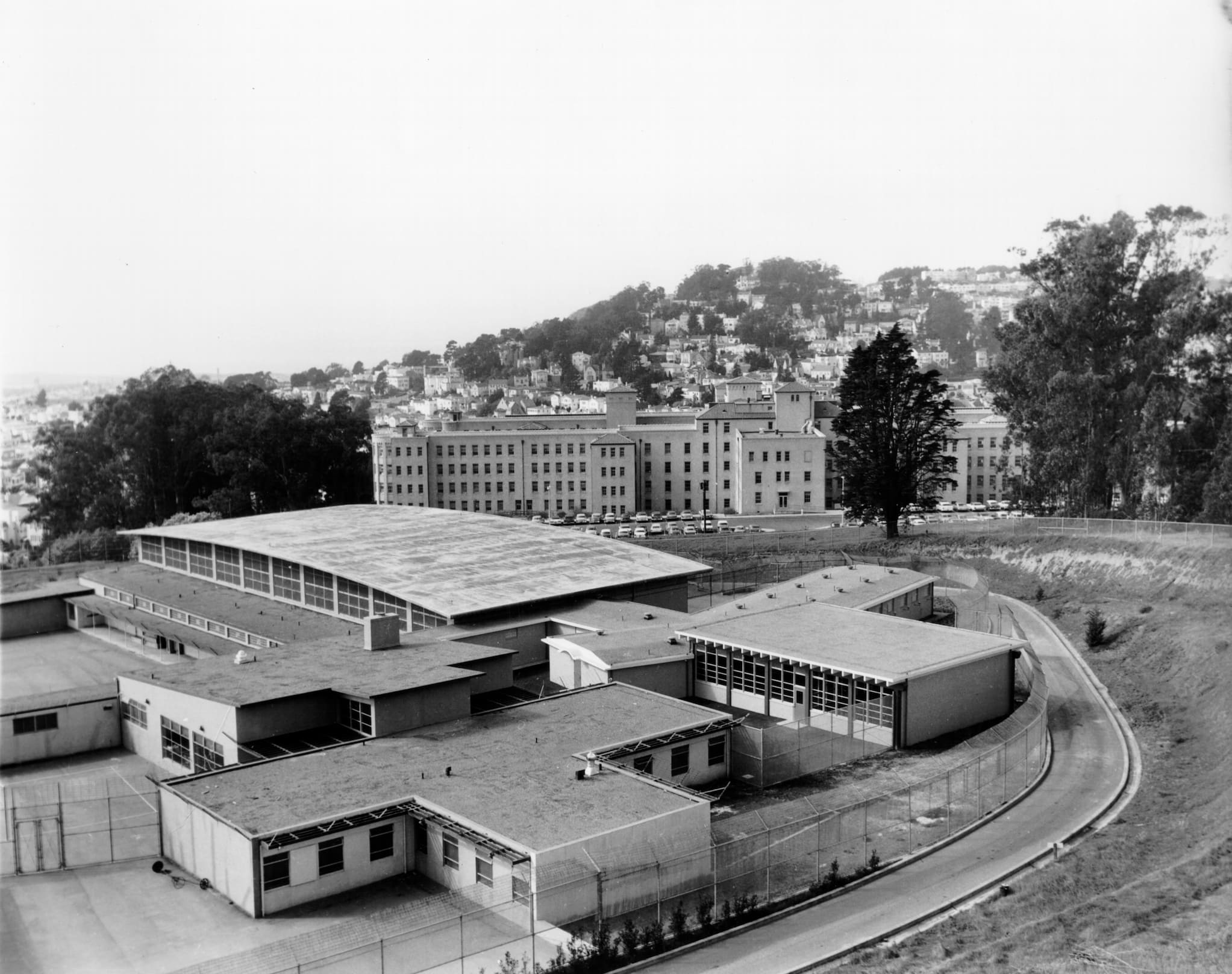 Aerial view of a maximum security cottage surrounded by barbed wire fencing. In the background is a city along a hill.