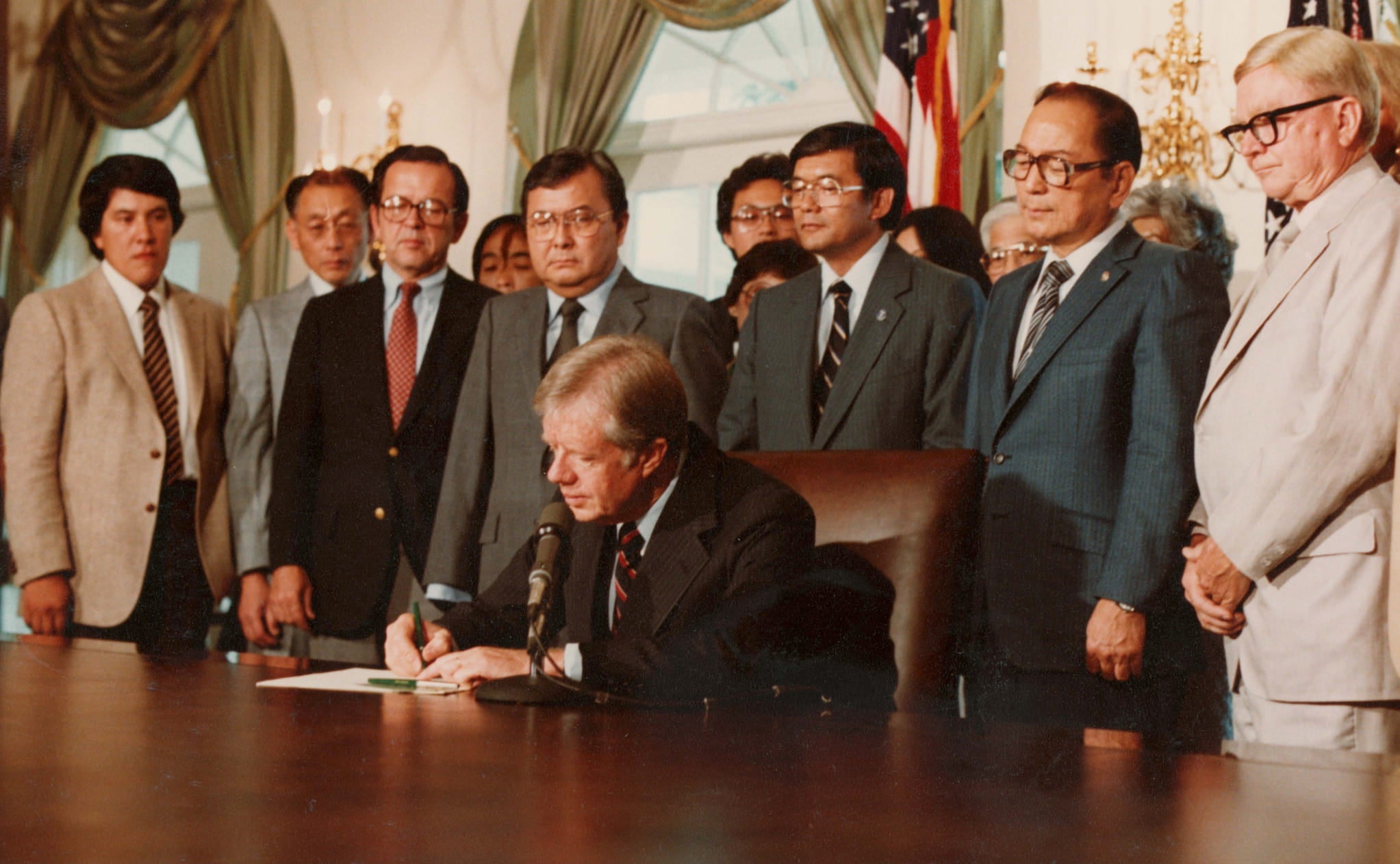 A group of Japanese men in suits stand behind a seated President Carter as he signs the Commission of Wartime Relocation and Internment of Citizens Act.