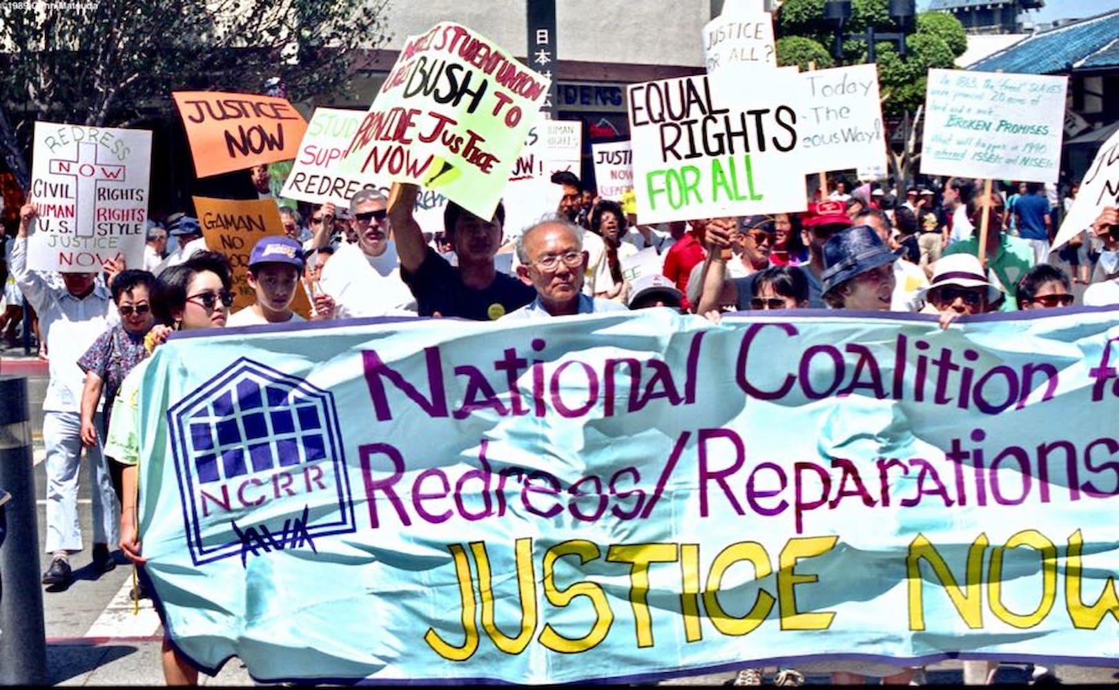 Individuals marching while holding signs and banner. A large banner in the front reads "National Coalition for Redress/Reparations Justice Now."