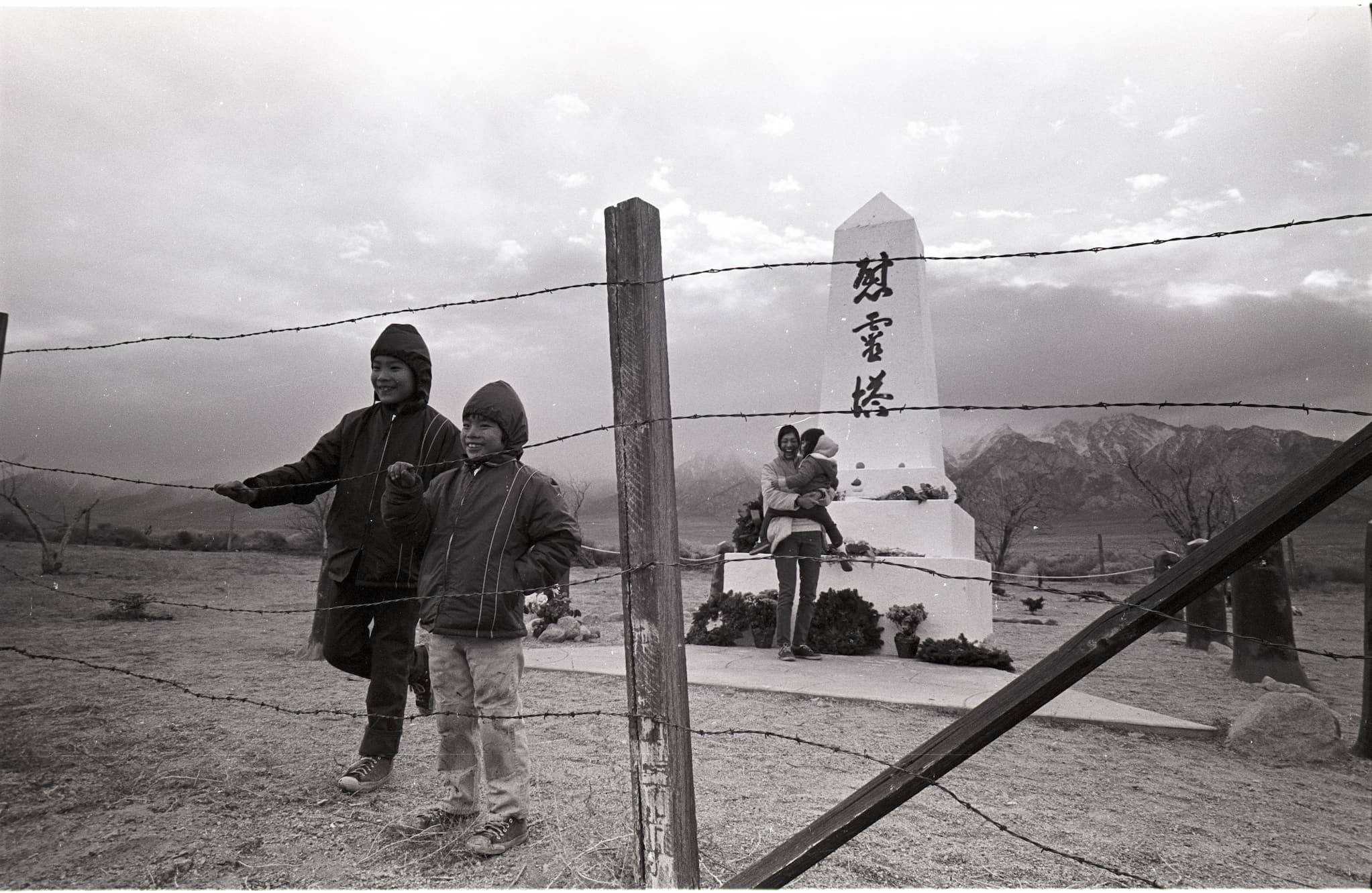 Two boys stand and hold onto a wired fence while their mother stands in the back holding a baby. Behind is the Manzanar Cemetery Monument.