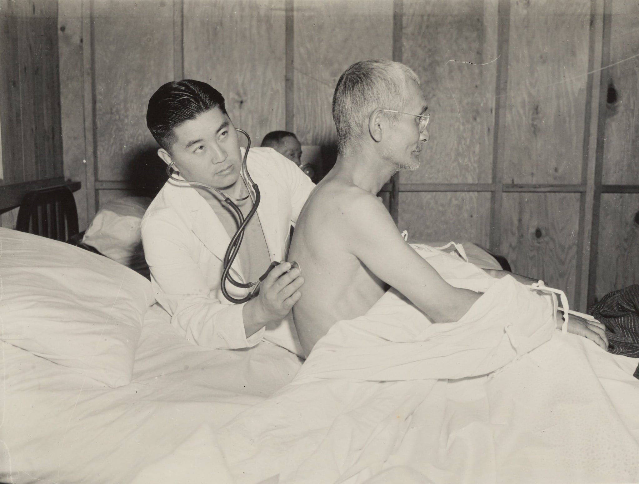 Japanese American doctor James Goto in white coat examines an elderly man in a bed by using a stethoscope on his back.