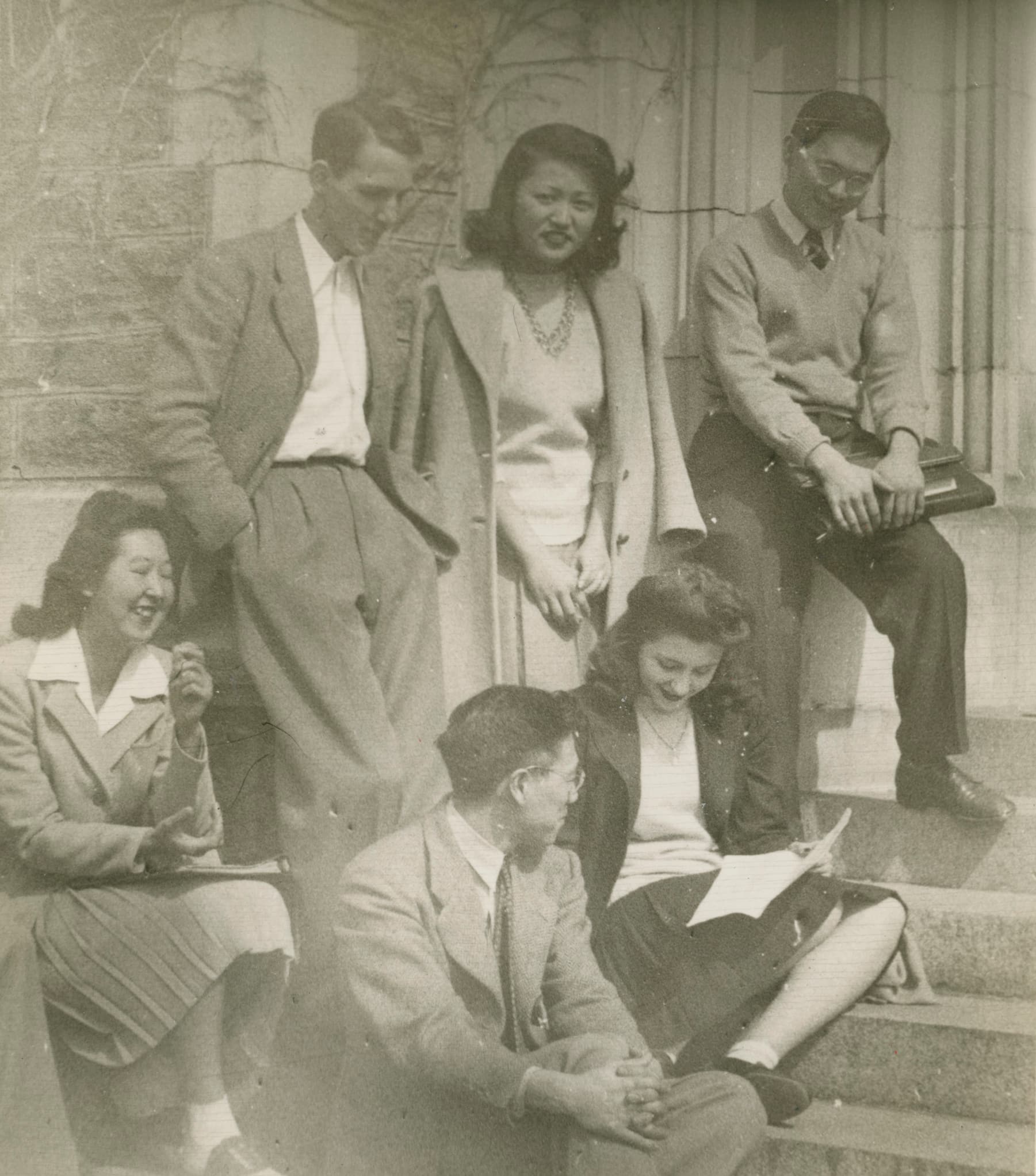 Four Nisei students dressed in formal attire sit and smile on the steps with two Caucasian people looking at papers.