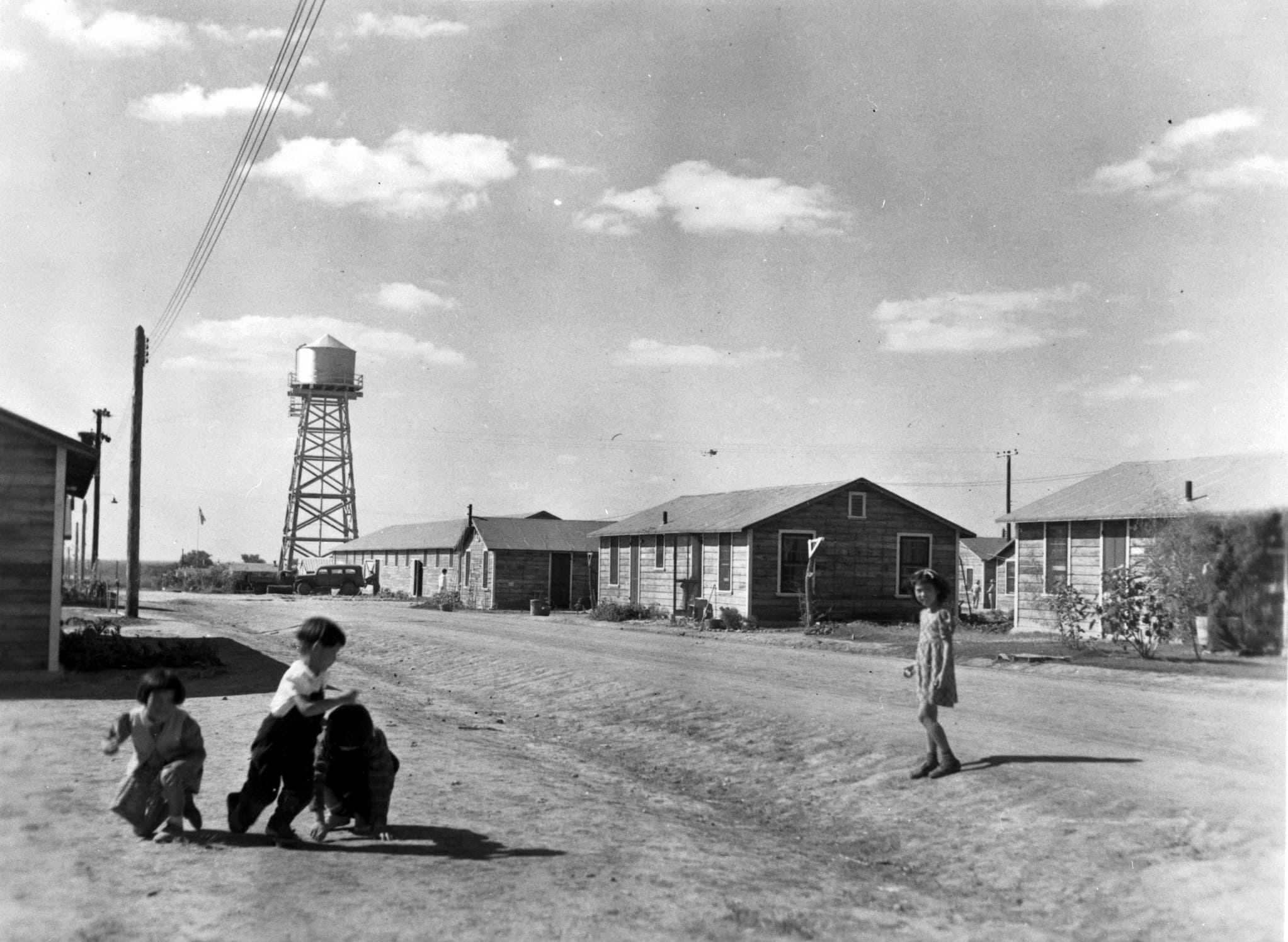 Three children playing outside in the forefront. In the background are a few buildings and a water tower on a barren land in Crystal City, Texas.