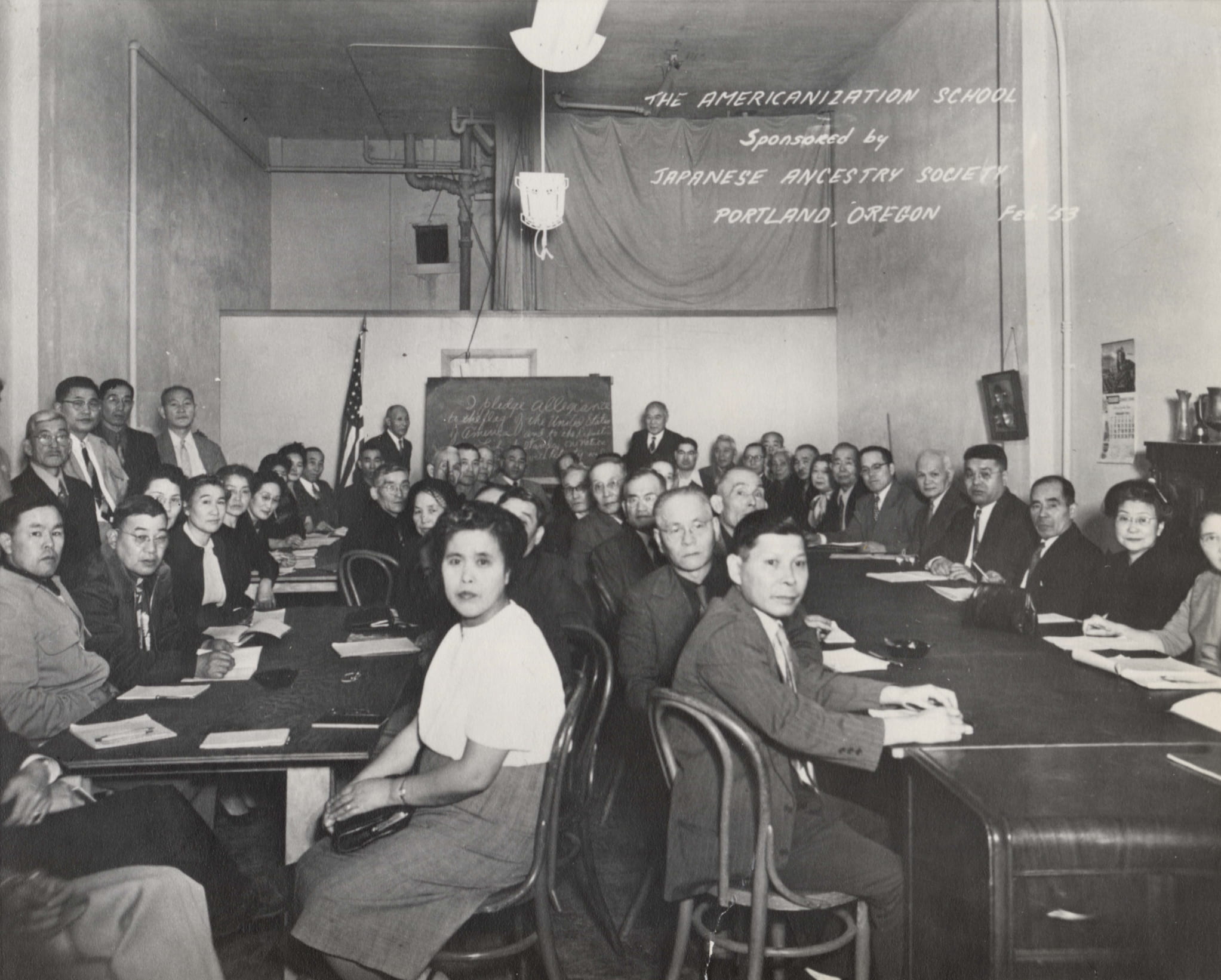 Black and white photo of two long tables in a classroom crowded with Japanese Americans in business clothing. Label reads "The Americanization School.