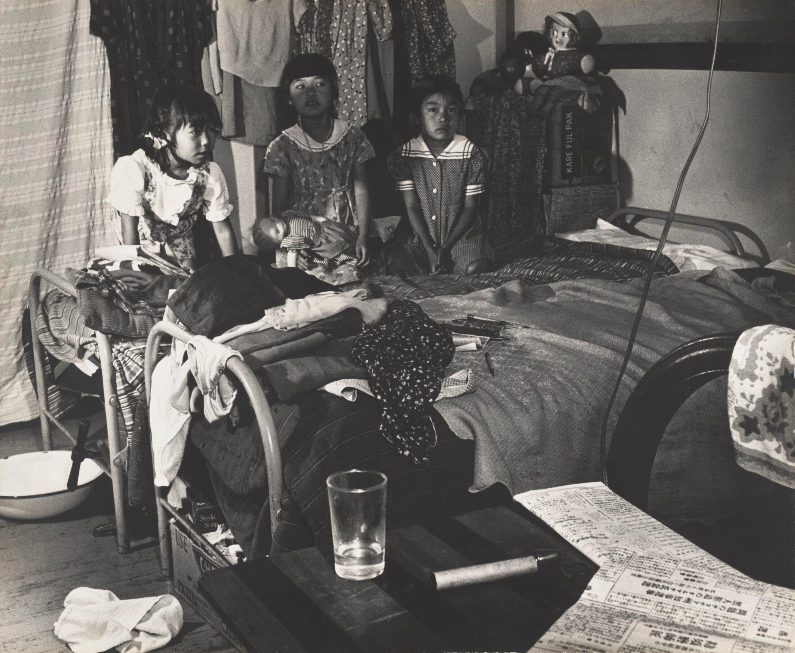 Three Japanese American girls sit behind two beds with clothes on top inside Evergreen Hostel. Center girl has a doll on her lap.