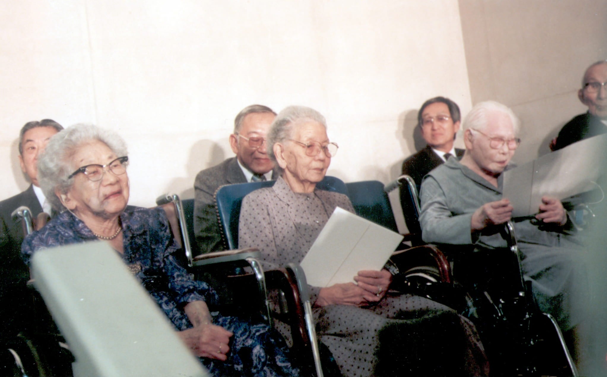 Three Issei women sitting in wheelchairs receive redress checks and formal apologies. Three Issei men sit behind.