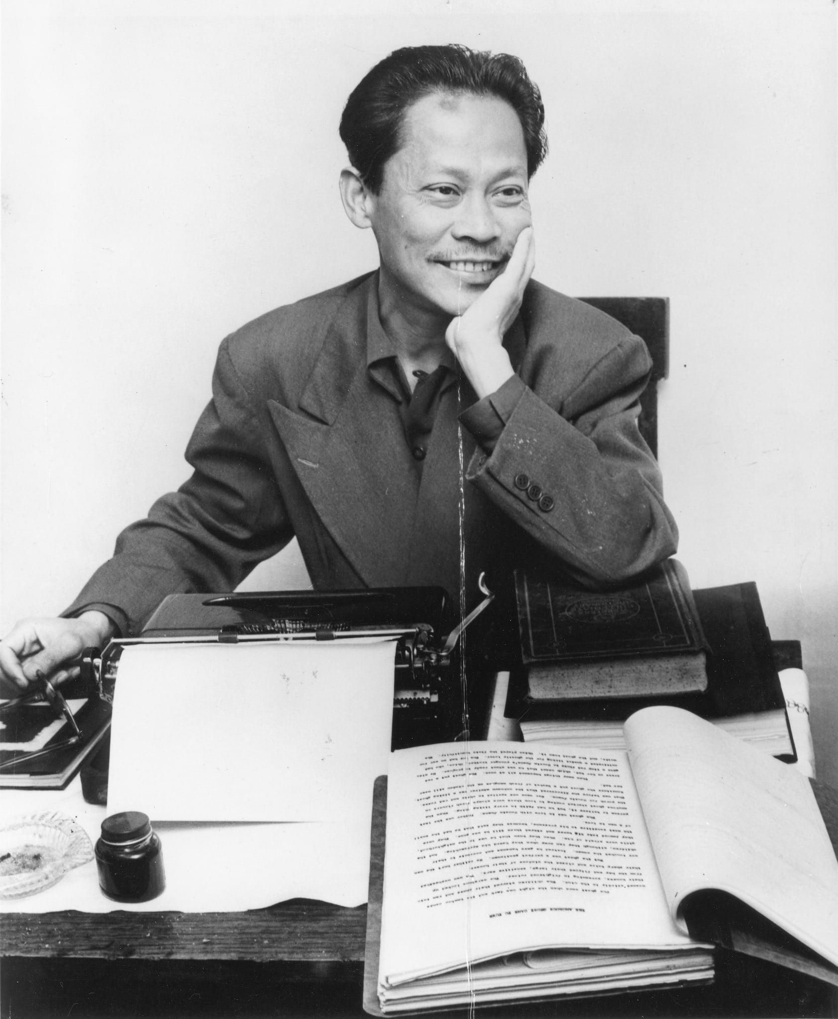 Filipino-American writer Carlos Bulosan sits at a writing desk with his hand on his cheek, smiling. A typewriter and books are visible on the desk.