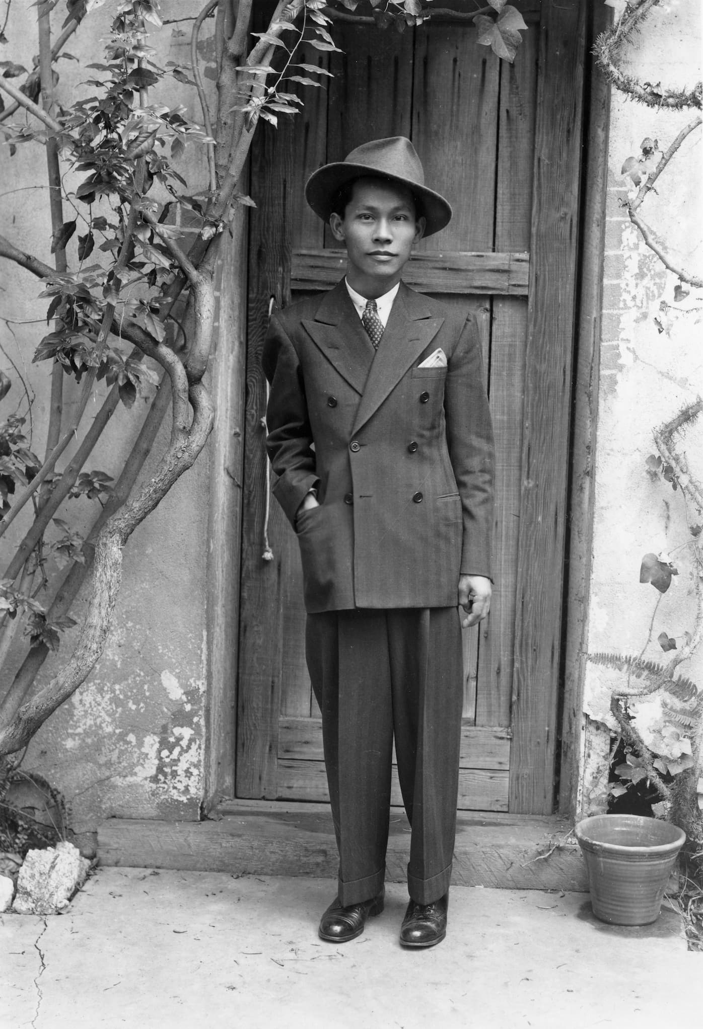 Filipino-American writer Carlos Bulosan stands in front of a wooden door wearing a full suit.