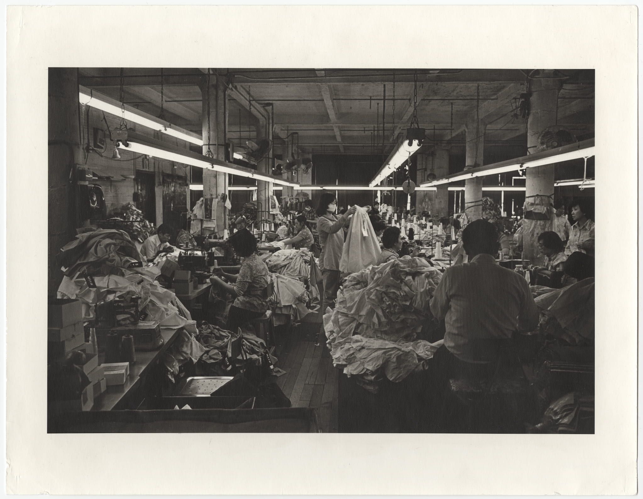 Interior view of crowded garment factory floor with workers surrounded by piles of clothes.