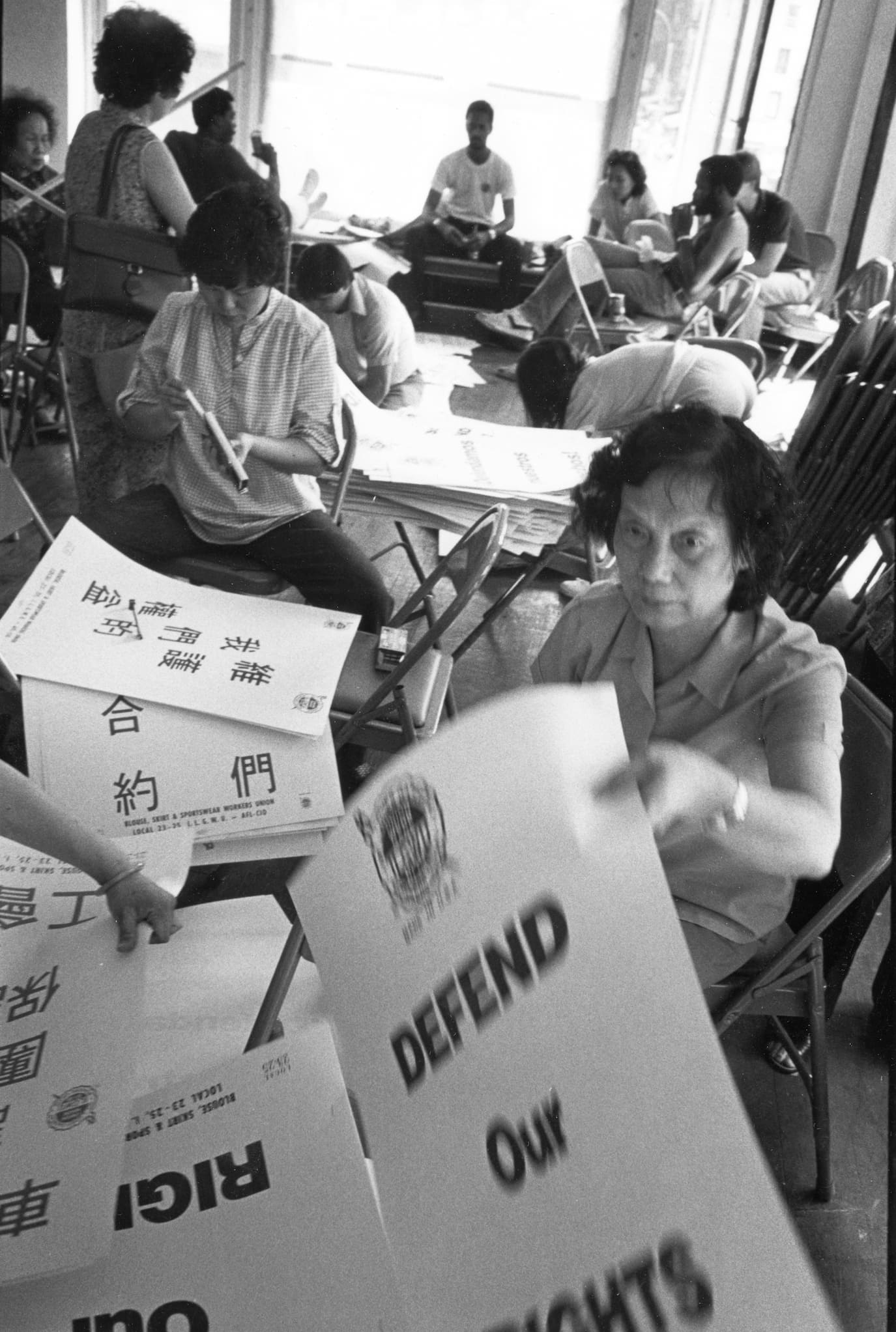 Volunteers, seated around tables, prepare signs in English and Chinese ahead of rally. One sign reads "Defend Our Union Contract."