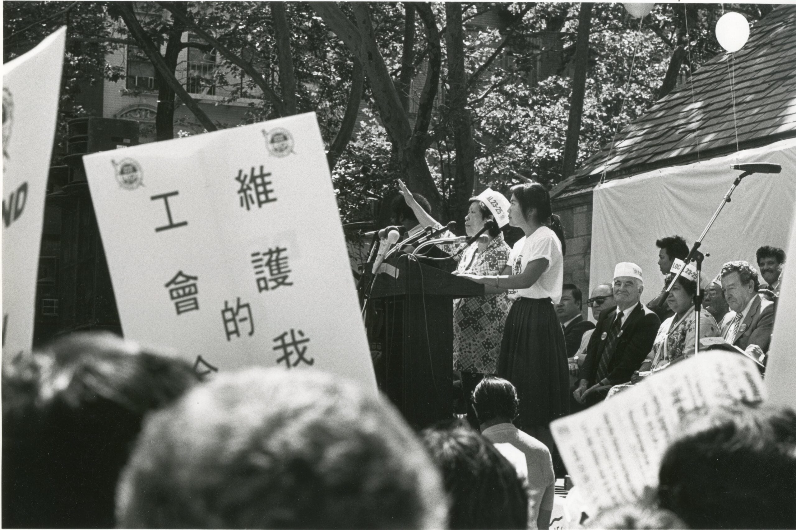 Woman with raised arm addresses crowd from podium at outdoor rally. Rally signs are in foreground.