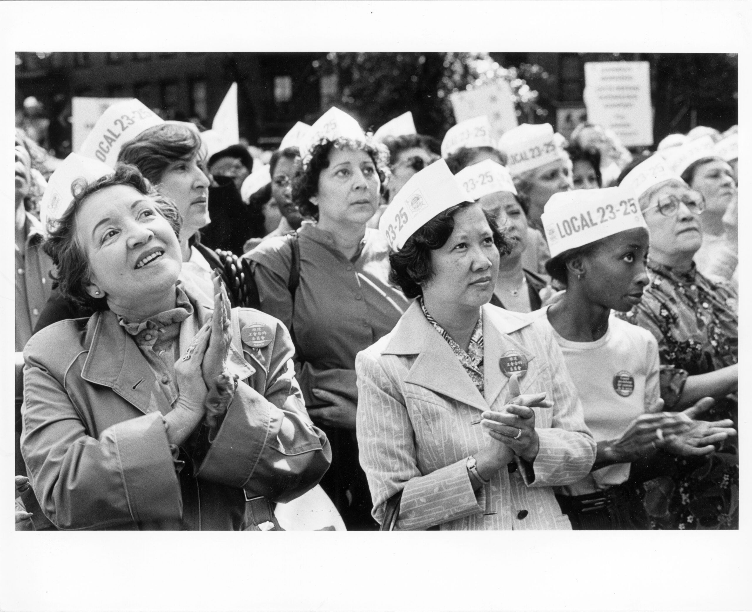 Group of women with "Local 23-25" hats listen to speakers at outdoor rally.